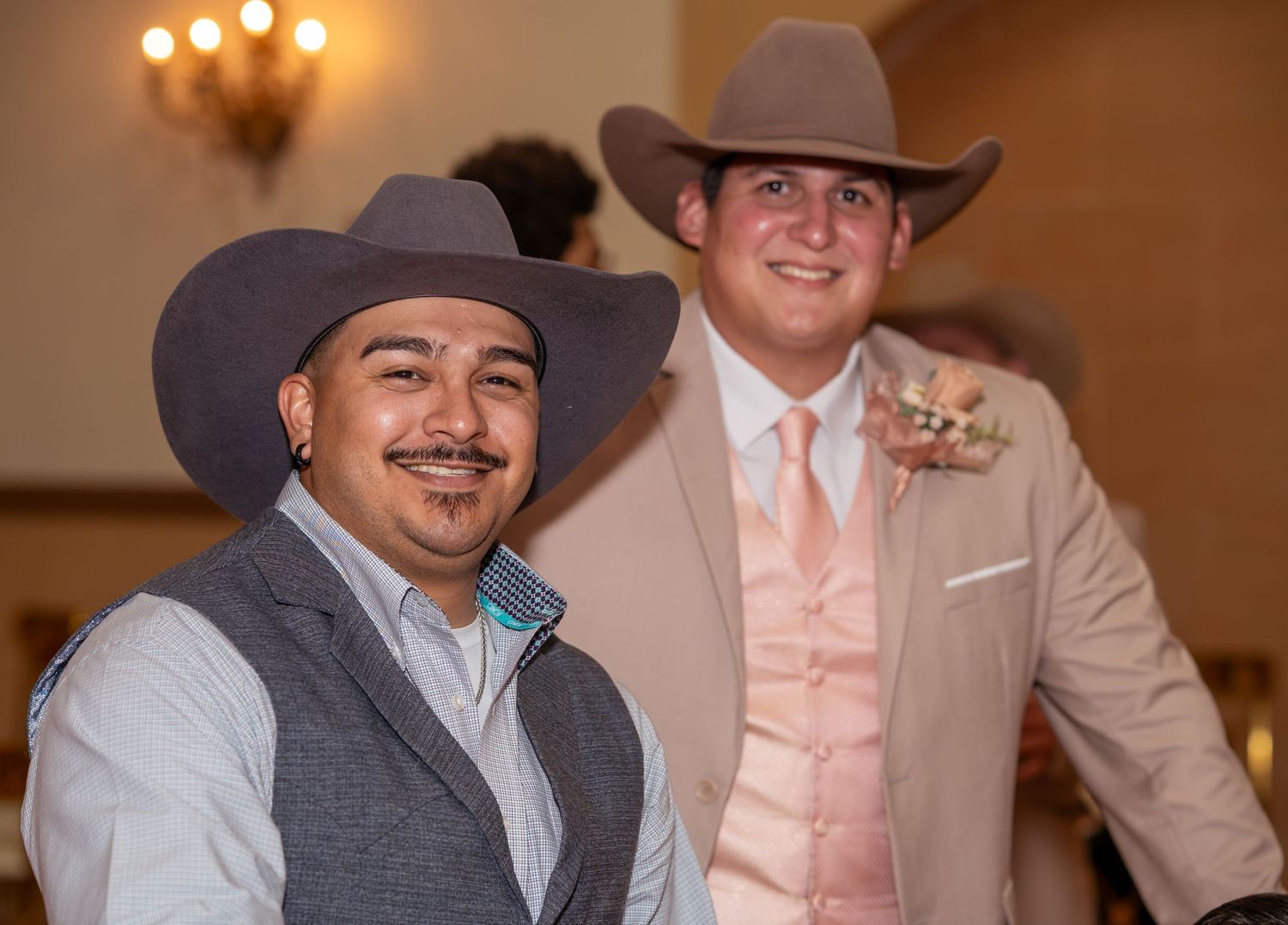 Two men in cowboy hats smile at a quinceañera celebration, one in a gray vest and one in a peach suit.