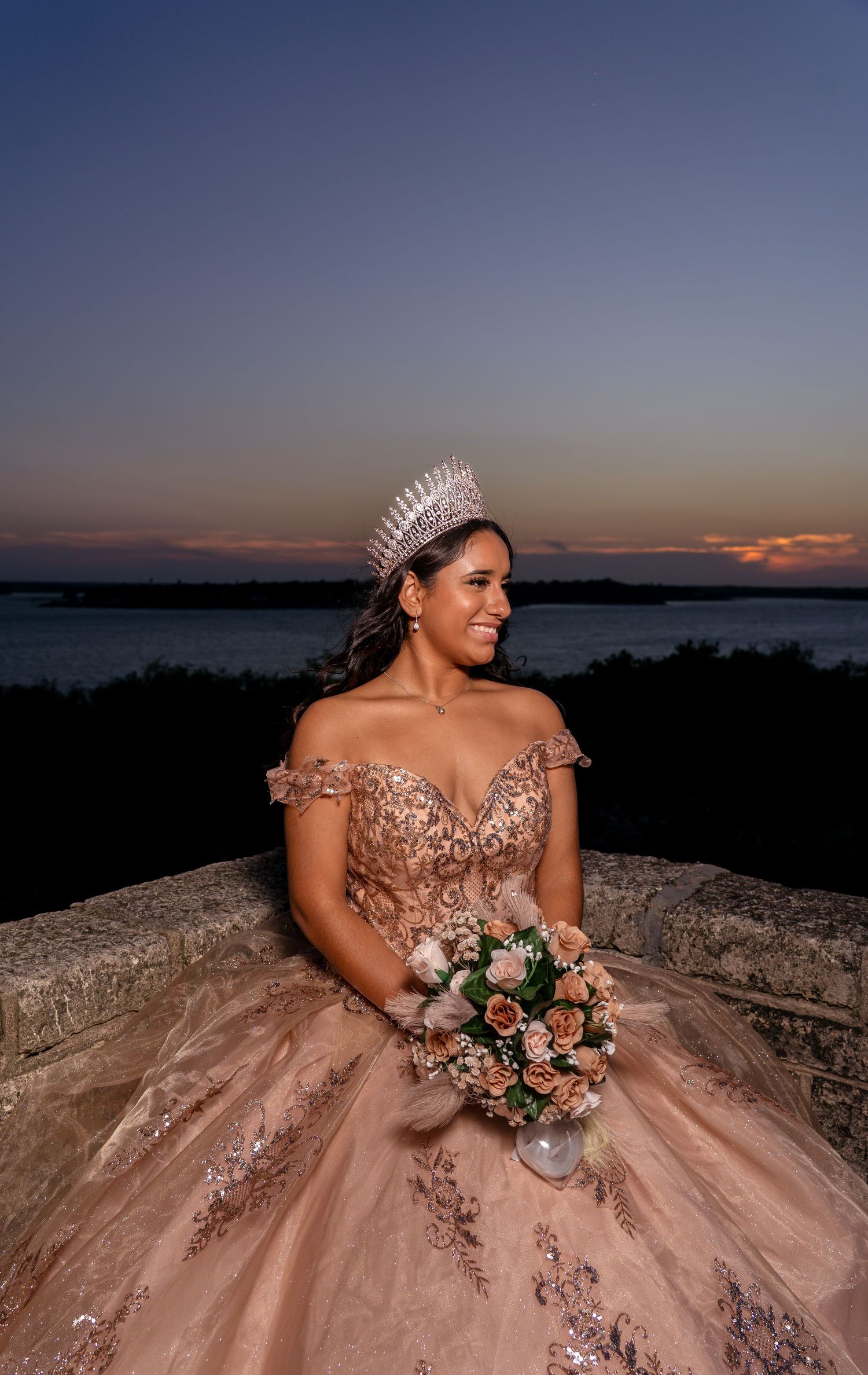 A bride in a sparkling rose gold gown with a crystal crown poses with her bouquet at sunset by the water.