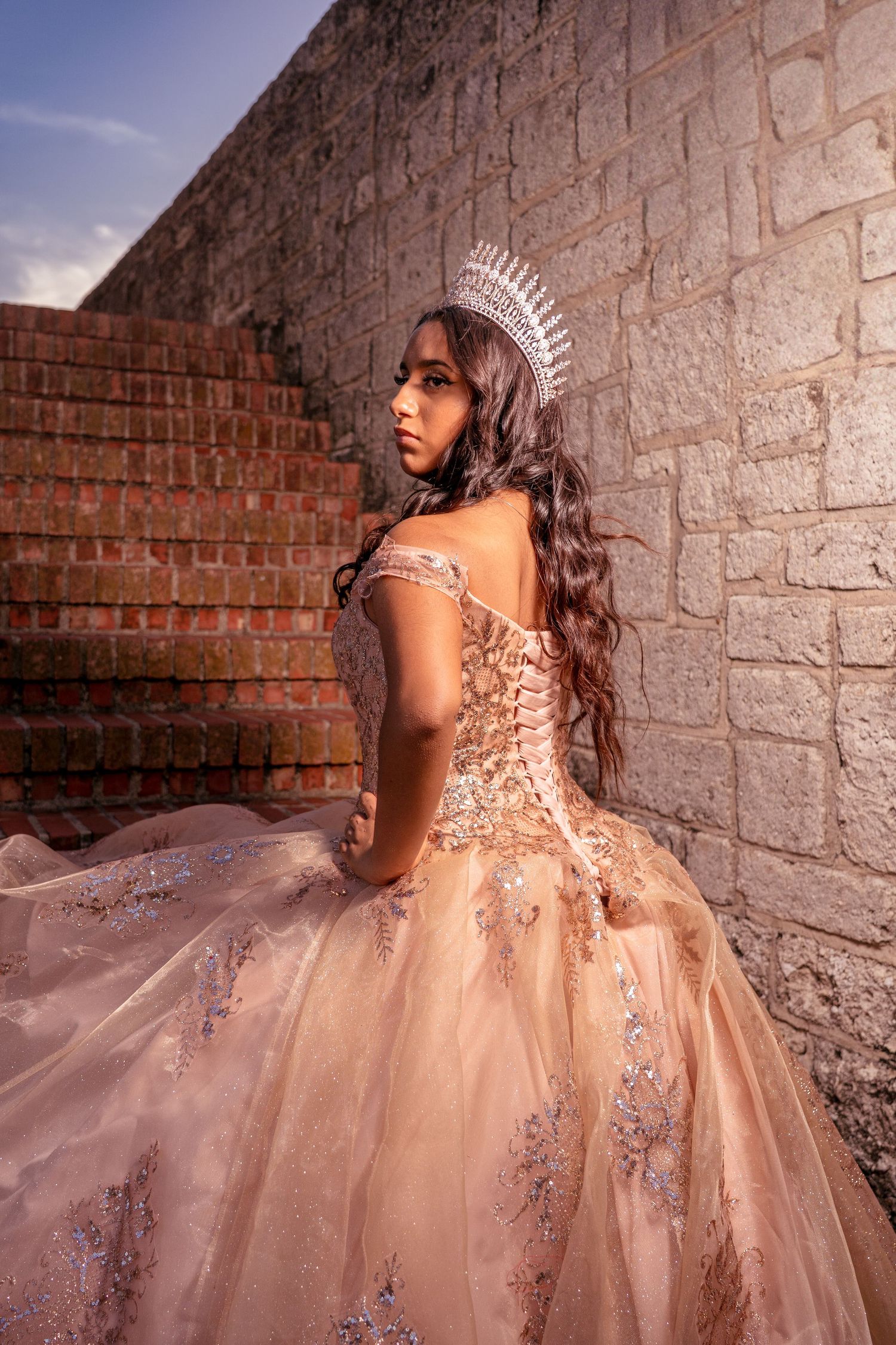 An elegant blush ball gown with corset back and crystal crown photographed against a stone fortress wall.