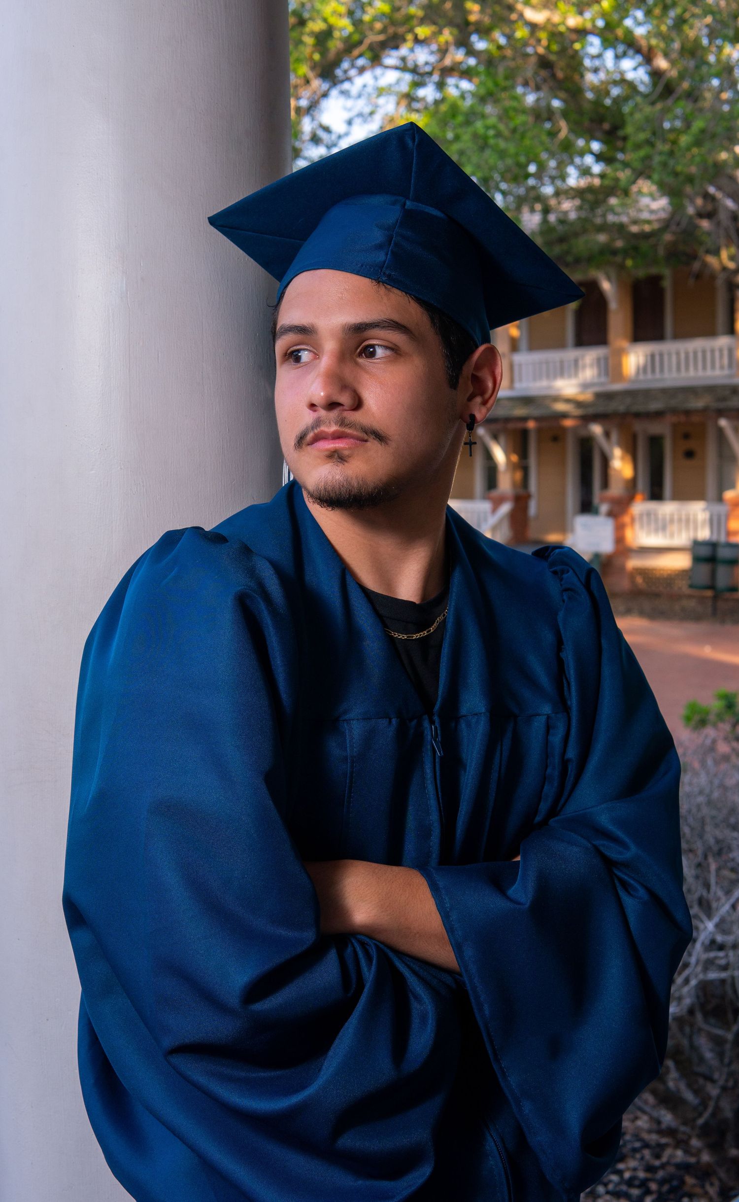 Graduate in navy blue cap and gown leans thoughtfully against white column during outdoor graduation photoshoot.