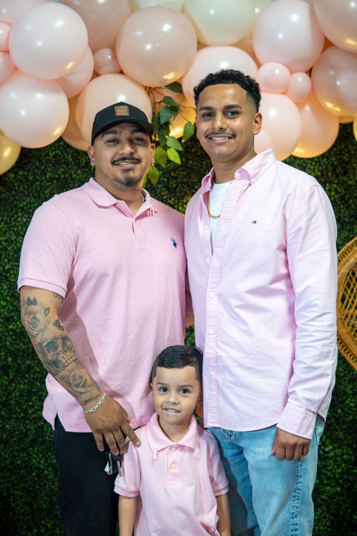 Family wearing matching pink polo shirts posed together in front of balloon arch and greenery backdrop at celebration.