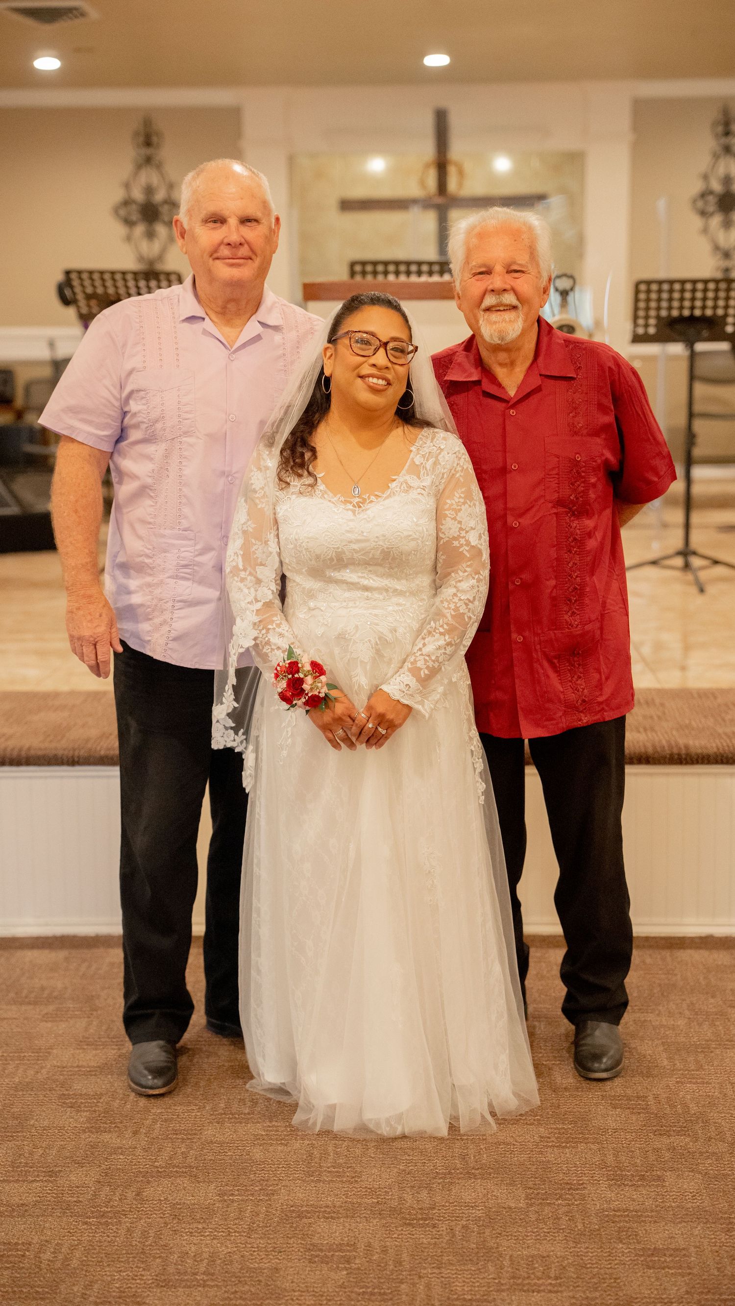 Bride in white lace gown with veil poses with two men in a church wedding venue.