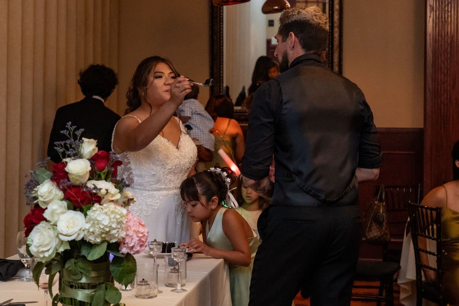 Bride and groom share a drink during wedding reception with floral centerpiece of red and white roses in foreground.
