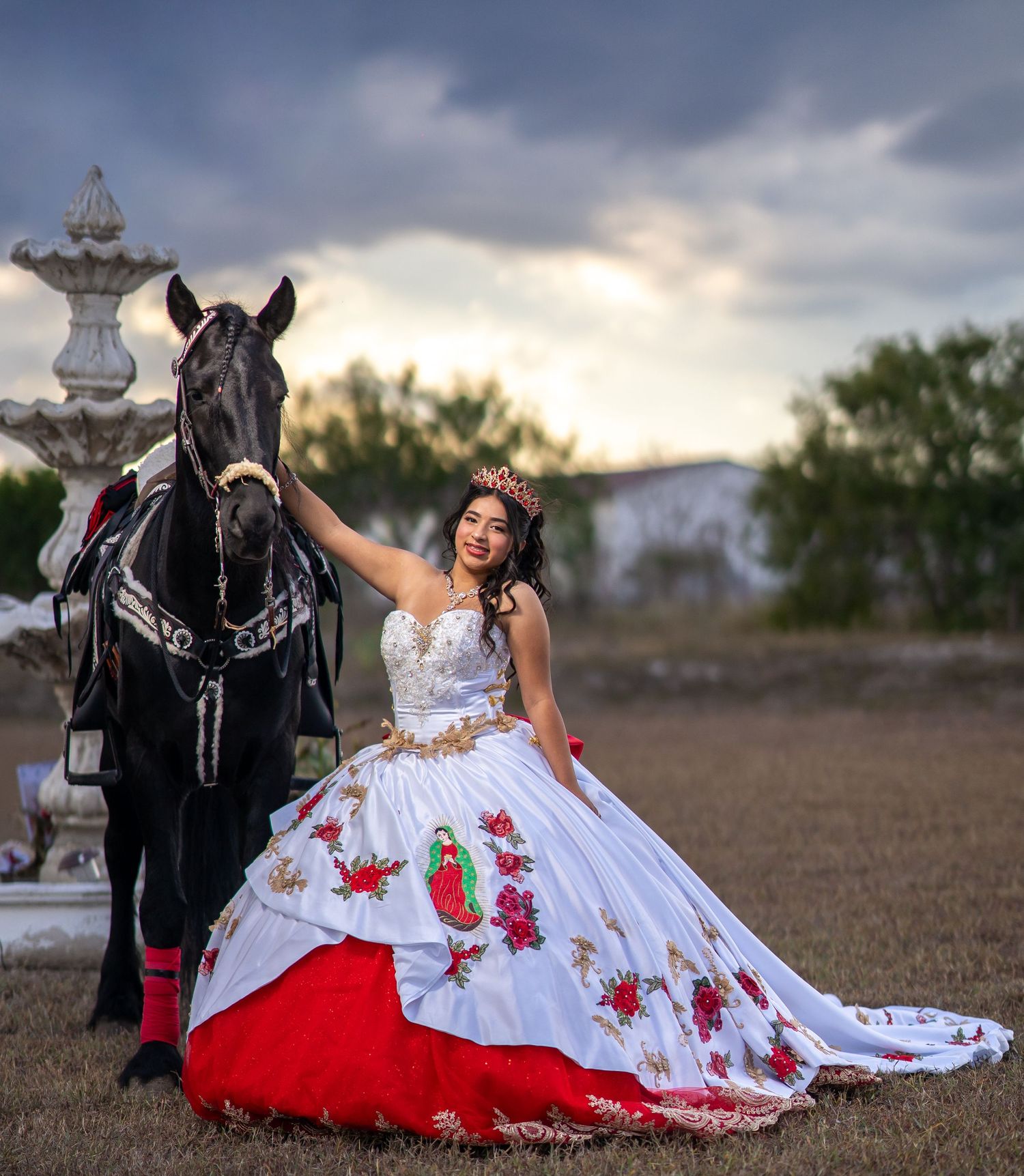 A bride in a white gown with red floral details poses with a black horse in a rustic outdoor setting at sunset.
