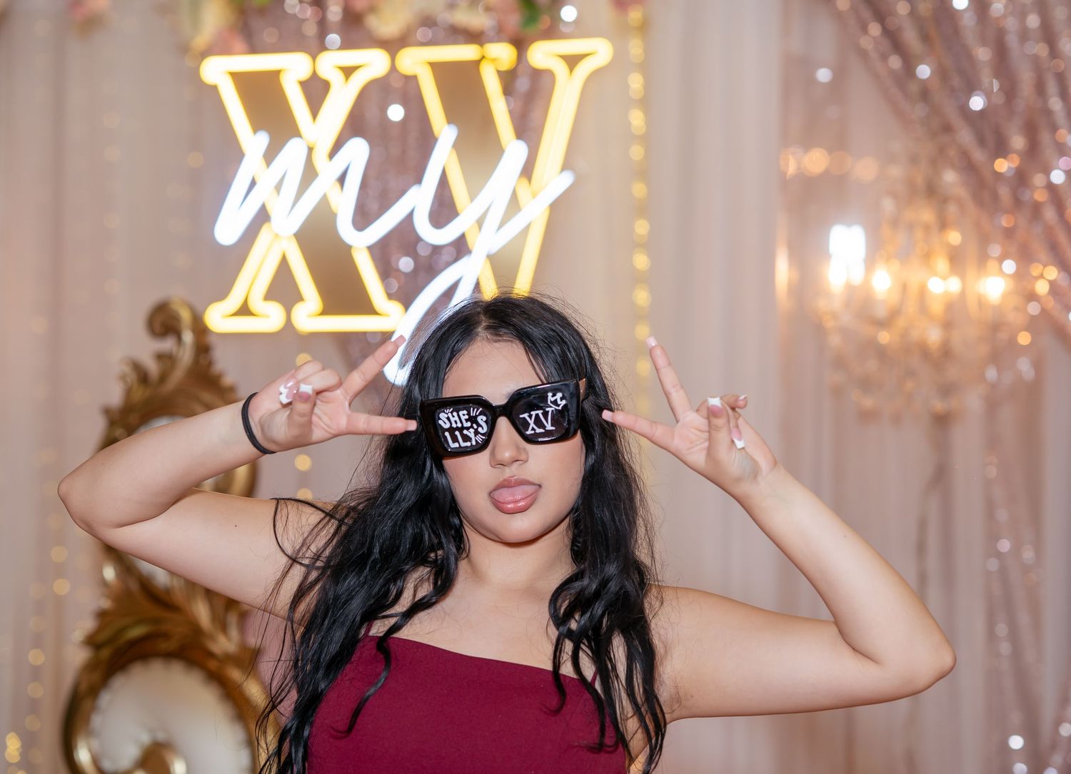 Young woman in burgundy dress poses with XV sunglasses at a quinceañera celebration.