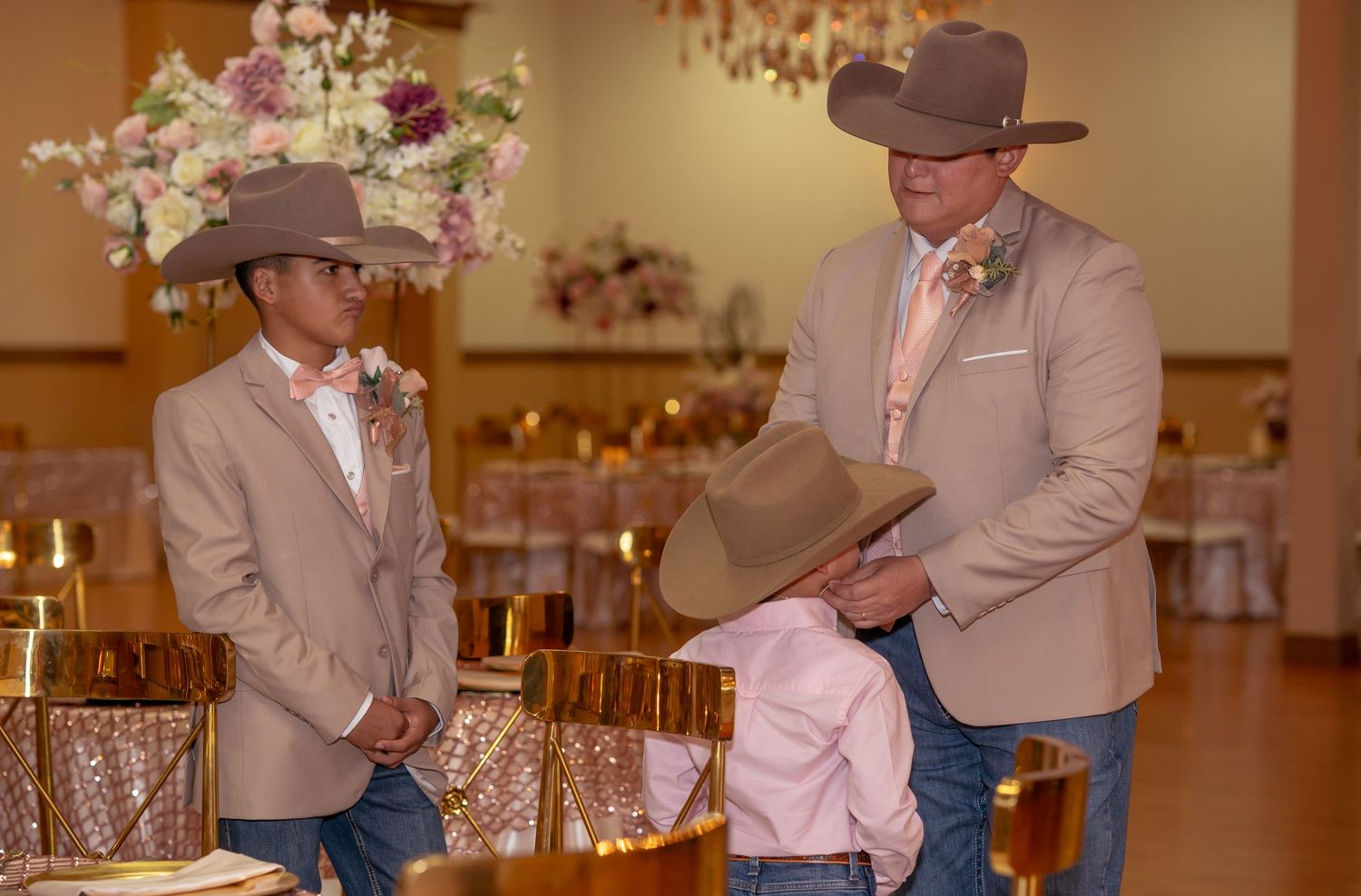 Man in tan suit and cowboy hat adjusting hat for young boy in matching outfit at a quinceañera celebration.