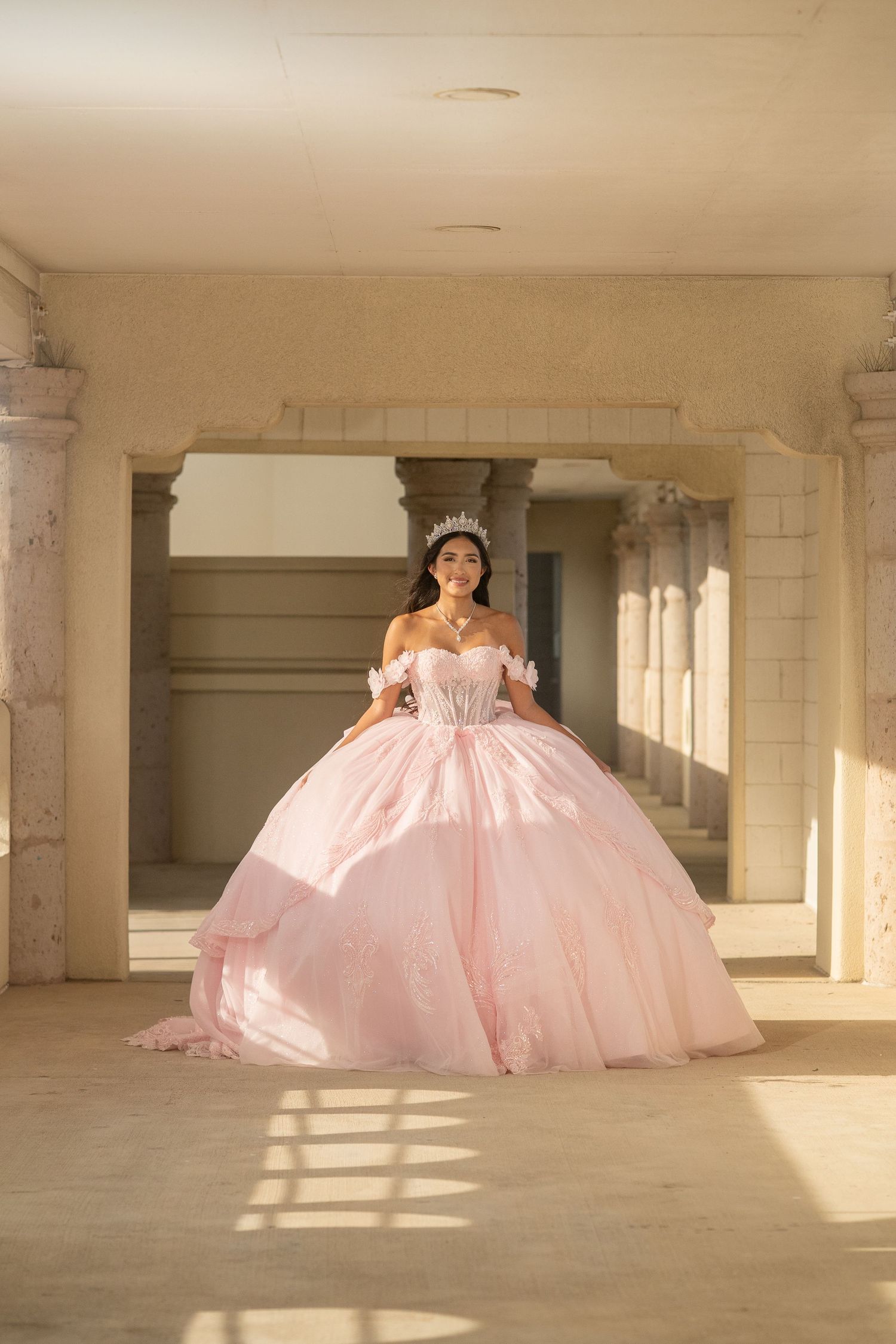 Young woman in a pink ball gown with tiara poses in a columned hall.