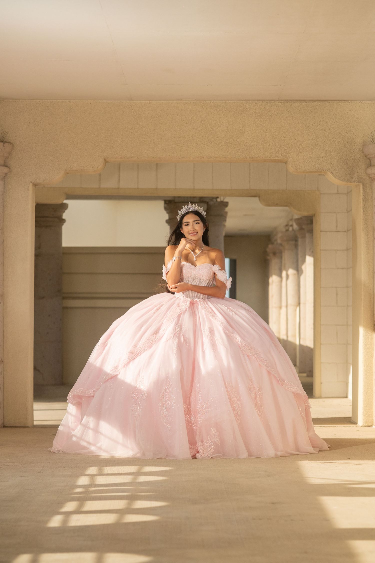 Quinceañera girl in voluminous pink ball gown and tiara poses under arched stone columns.