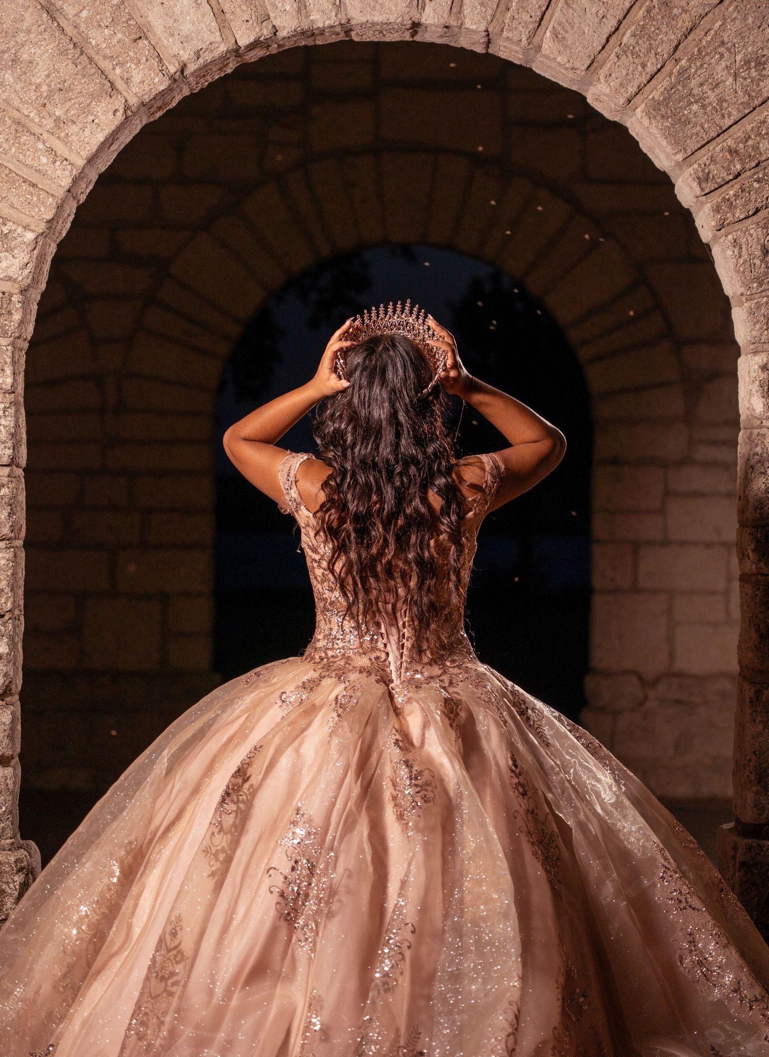 A back view of an ornate rose gold ball gown showcasing intricate beadwork under a stone archway.