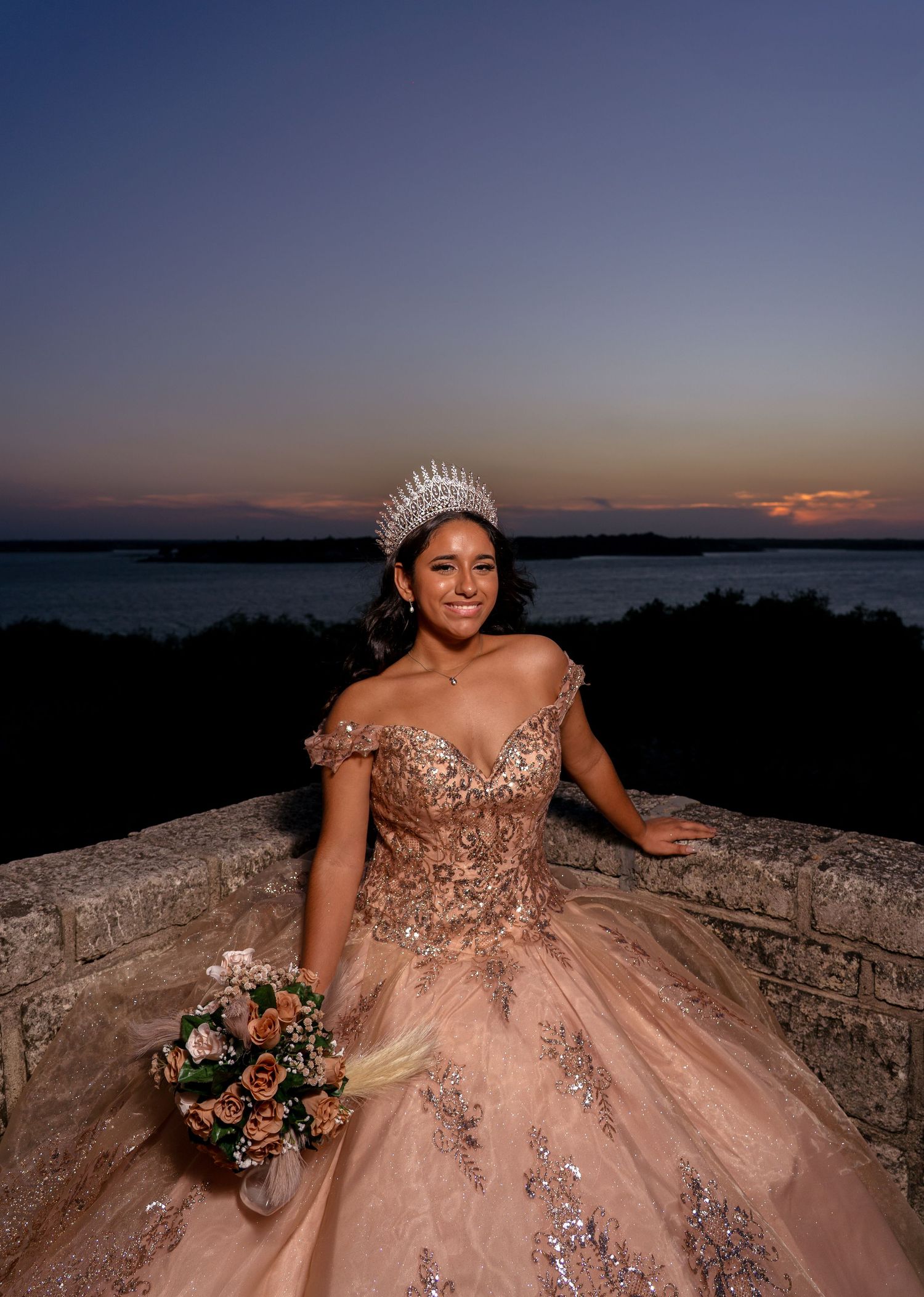 Beautiful evening portrait in a sparkly blush pink ball gown with tiara and bouquet against a sunset lakeside backdrop.