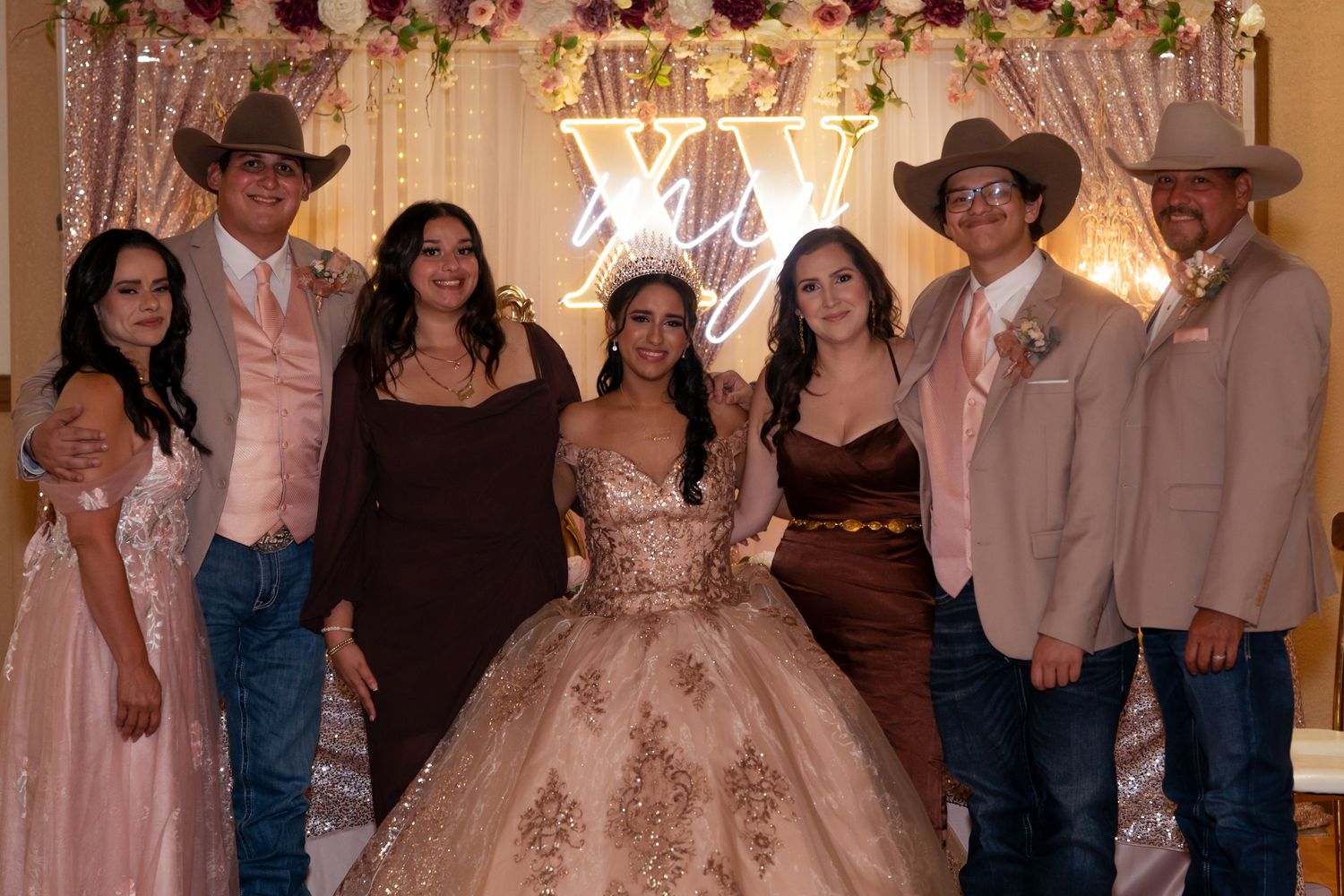 A Quinceañera in a gold ballgown poses with six family members in western attire before an XV illuminated backdrop.