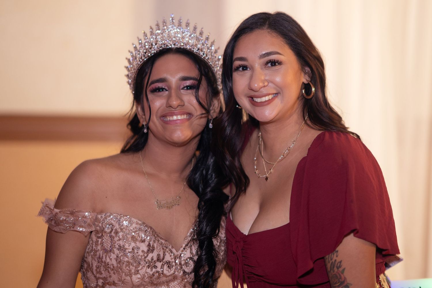 Quinceañera in gold gown and crystal crown smiles with a guest in red at her celebration.