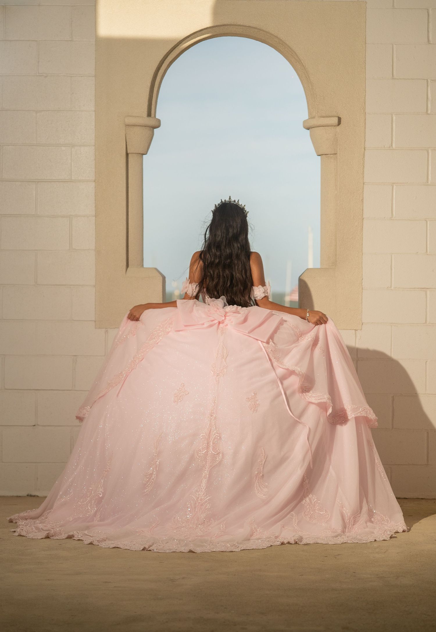 Back view of quinceañera in pink ball gown standing before stone arch overlooking ocean view.