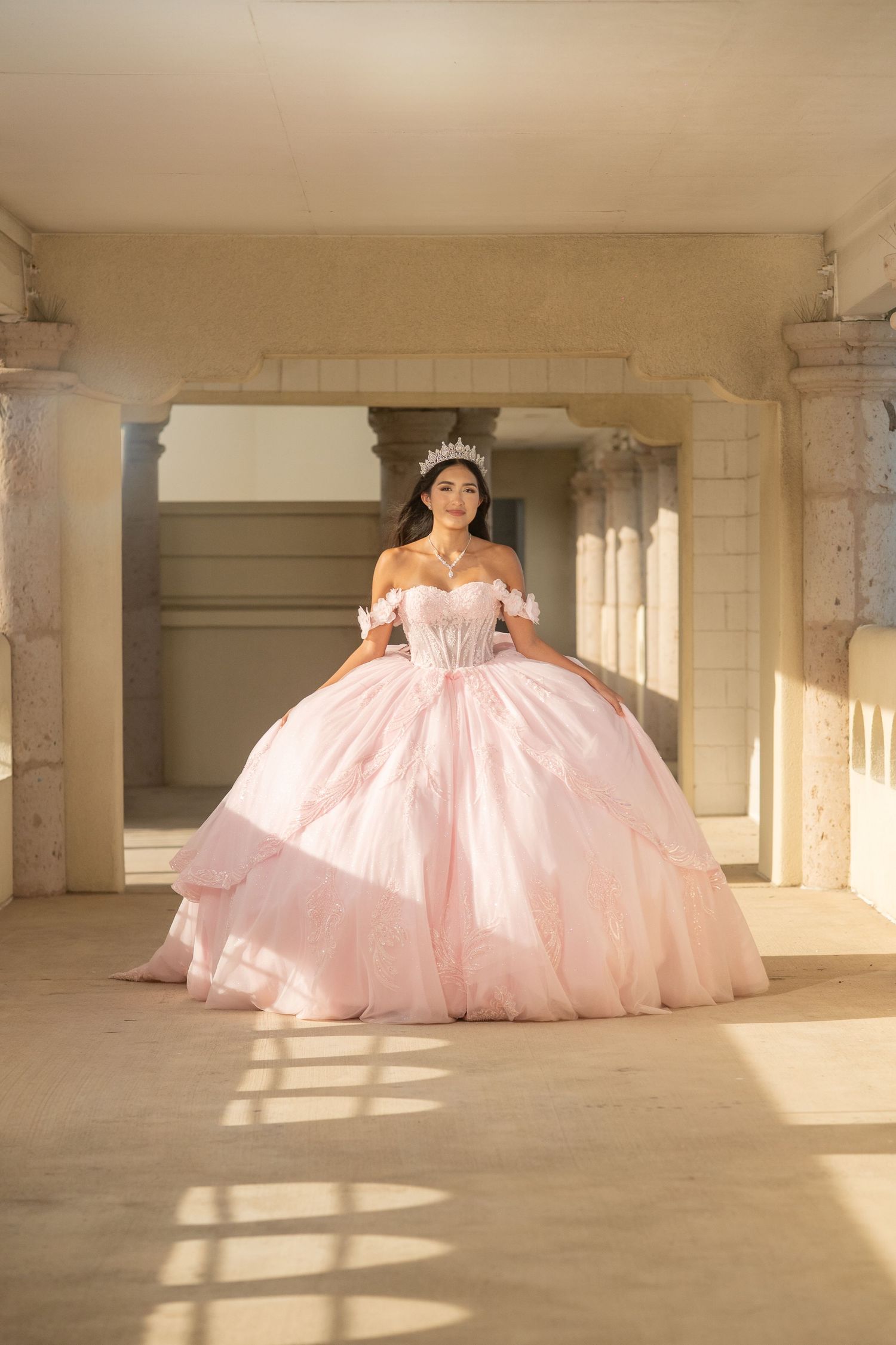 Young woman in a blush pink off-shoulder quinceañera ball gown and crystal tiara posing in a sunlit archway.