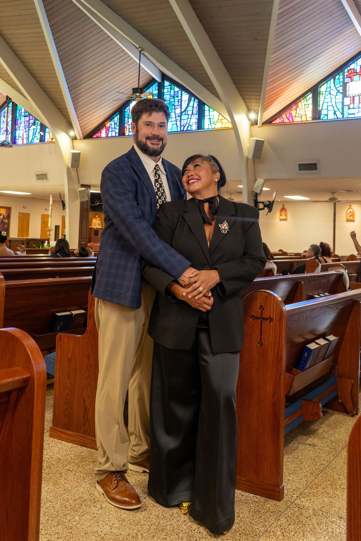 Couple poses together smiling in church with stained glass windows behind them.