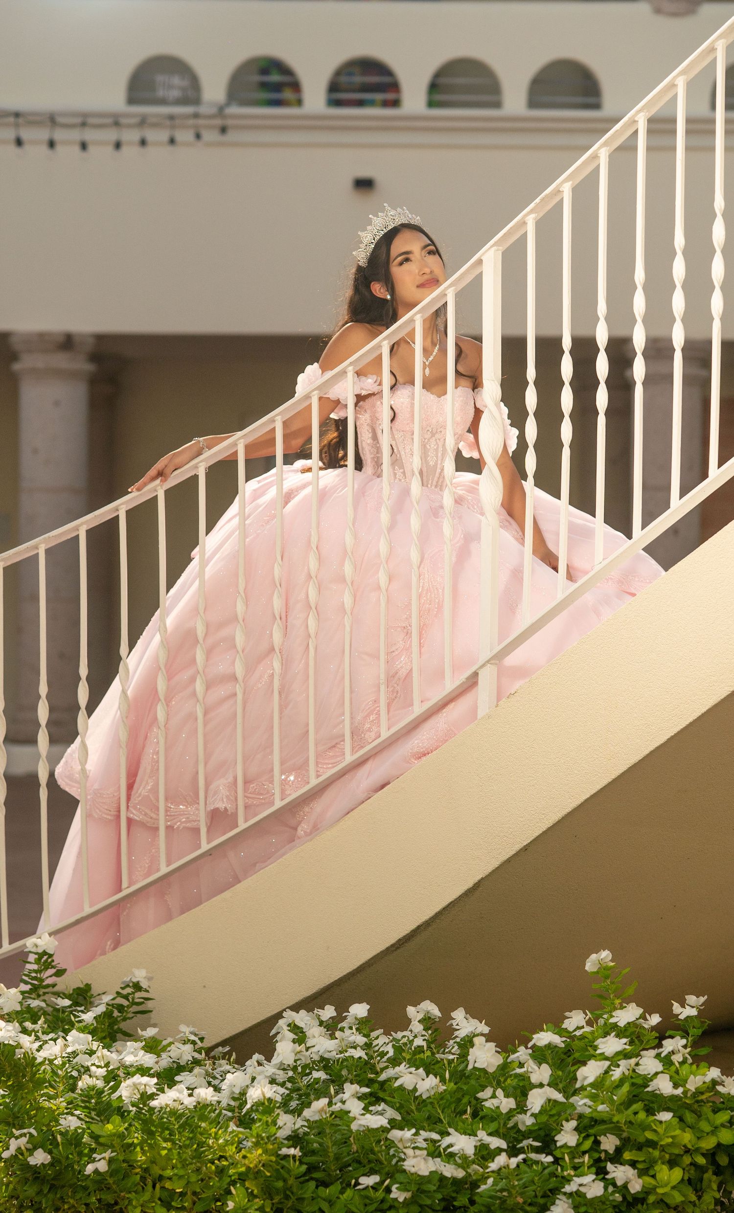 Young woman in pink ball gown and tiara poses gracefully on elegant curved staircase during quinceañera celebration.