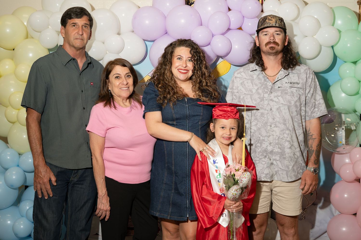Family poses with young child in red graduation cap and gown in front of colorful balloon backdrop.