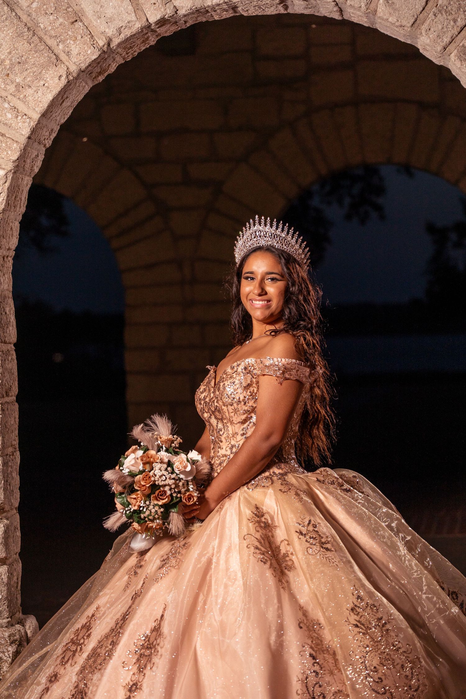 Rose gold quinceañera dress with crystal embellishments photographed in archway during dusk.