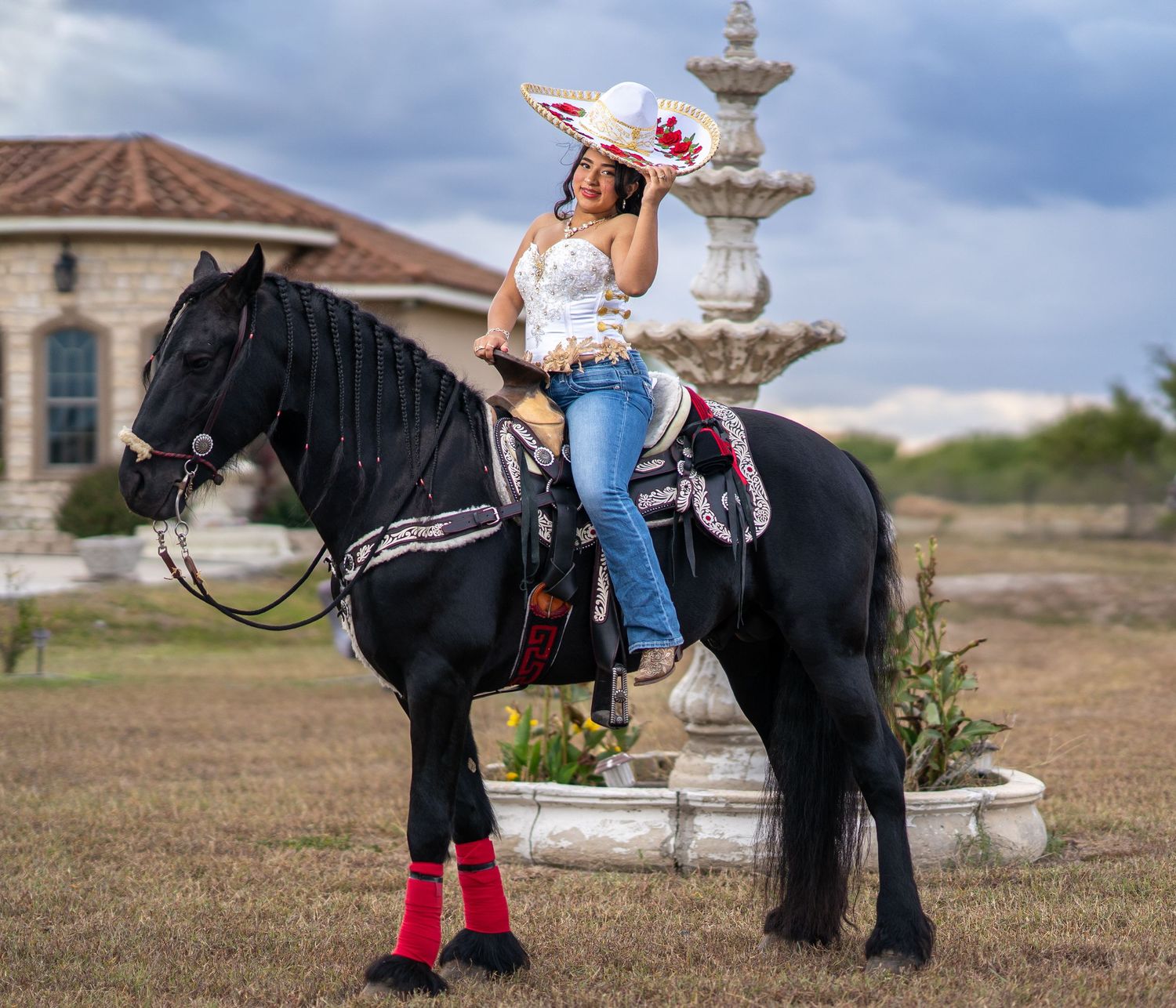 A rider in jeans and boots sits atop a majestic black horse near an ornate stone fountain and Mediterranean-style building.
