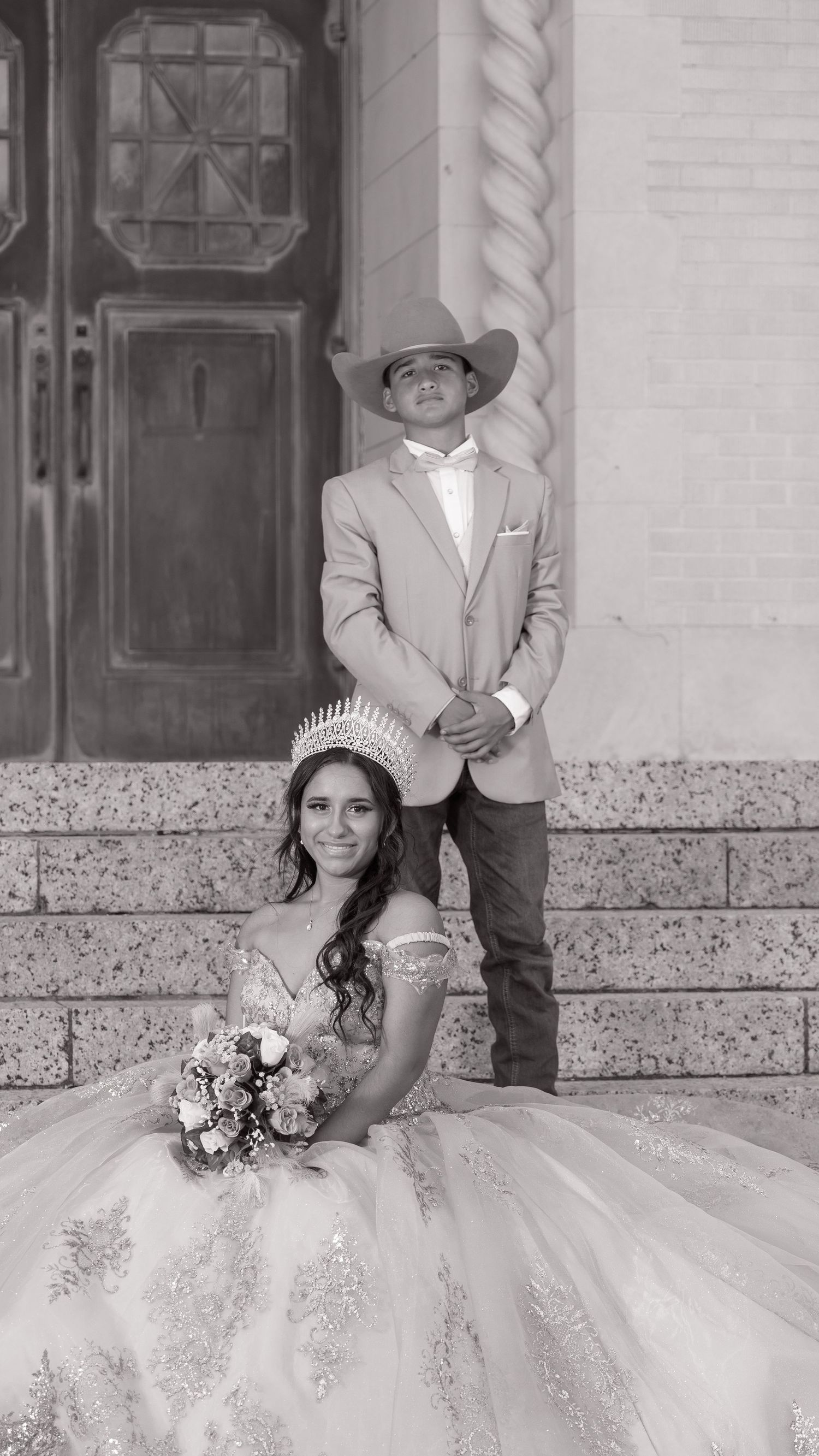 A bride in an ornate white ball gown sits on stone steps while someone in a light suit stands beside her.
