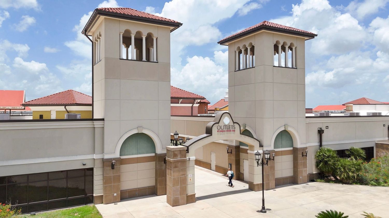 A panoramic view shows a beige Mediterranean-style building with arched windows and red-tiled roofs against a cloudy blue sky.