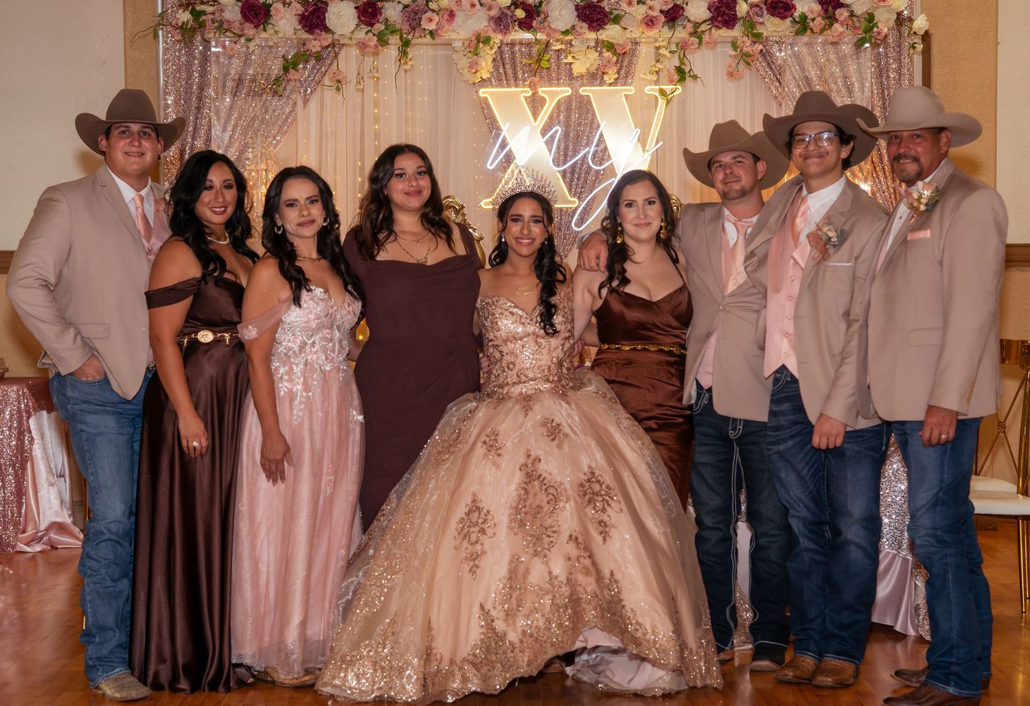 Group of eight people posing at a quinceañera celebration with XV decorations and floral backdrop.