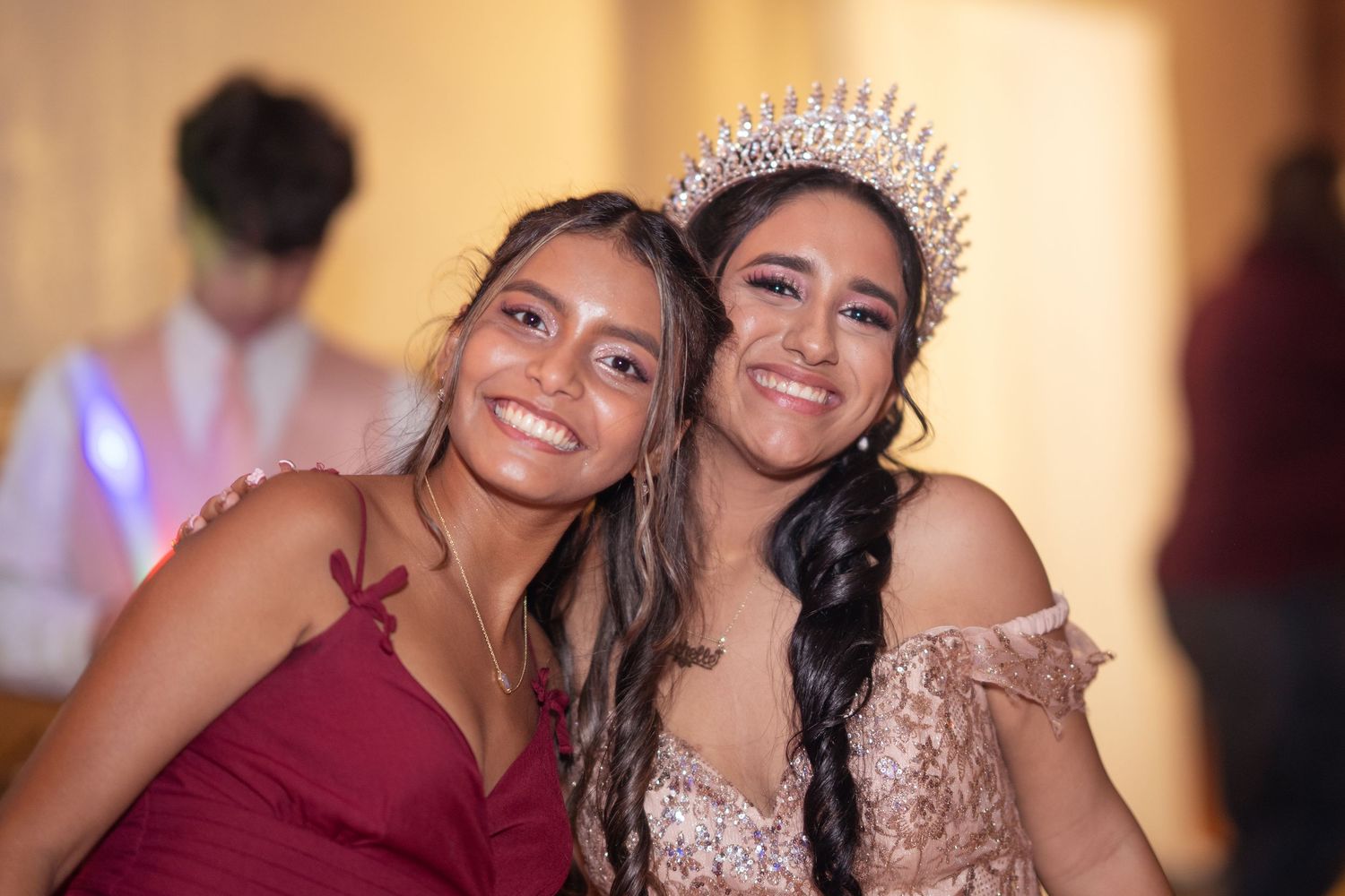Two young women smile together at a quinceañera, one wearing a sparkling crown and off-shoulder gown.