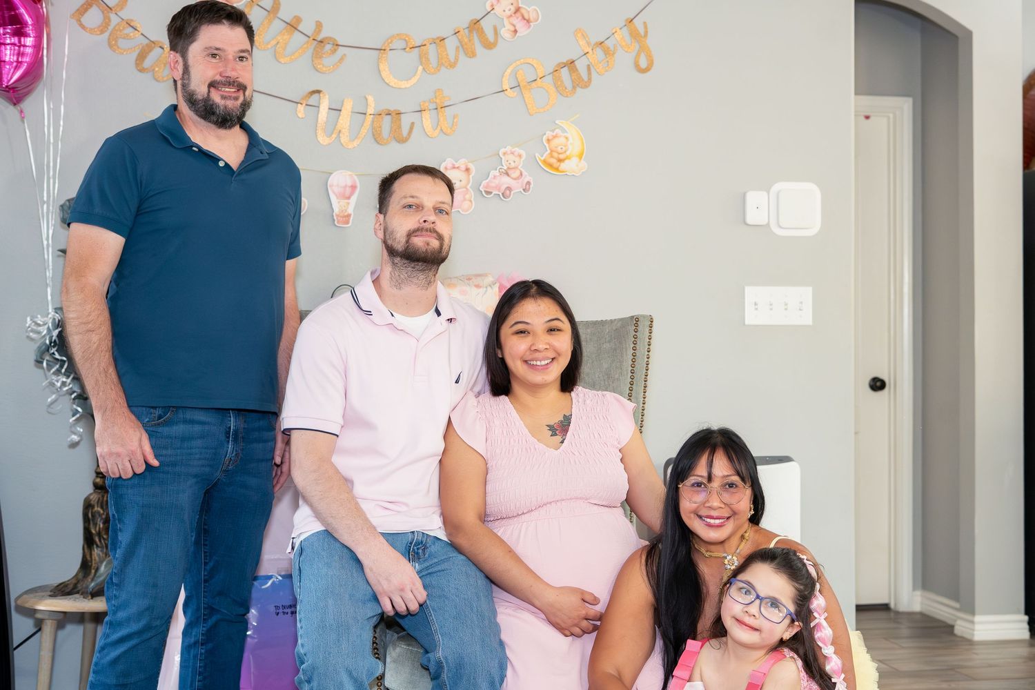 Happy family group posing together at a baby shower with pink balloons and decorative banner in a living room.