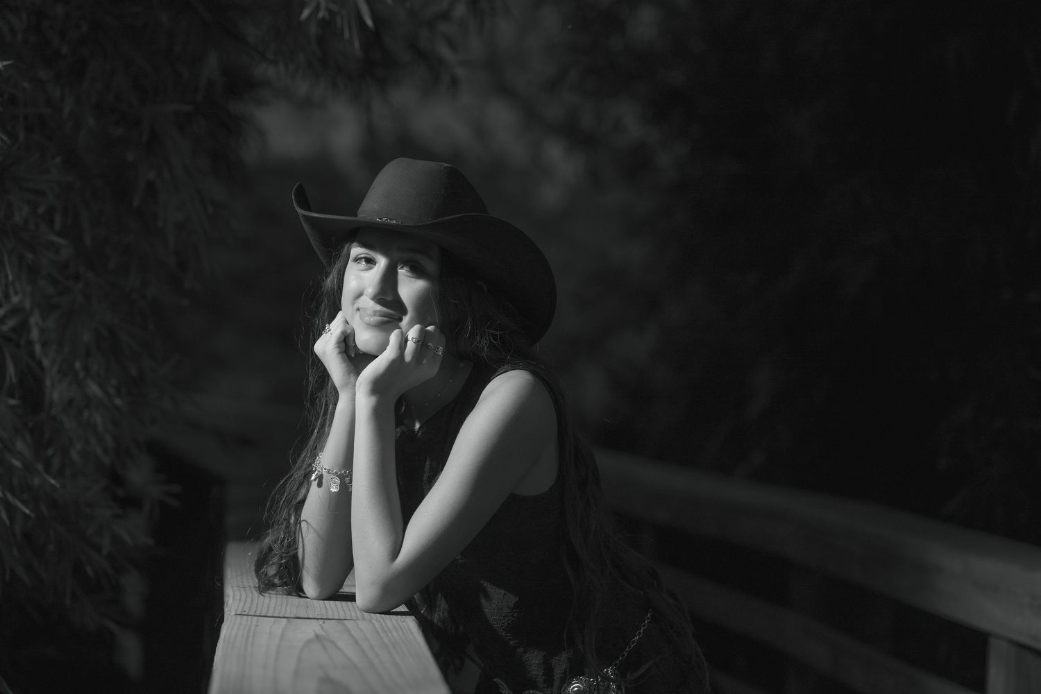 Black and white portrait of a smiling woman in a cowboy hat leaning on a wooden railing outdoors.
