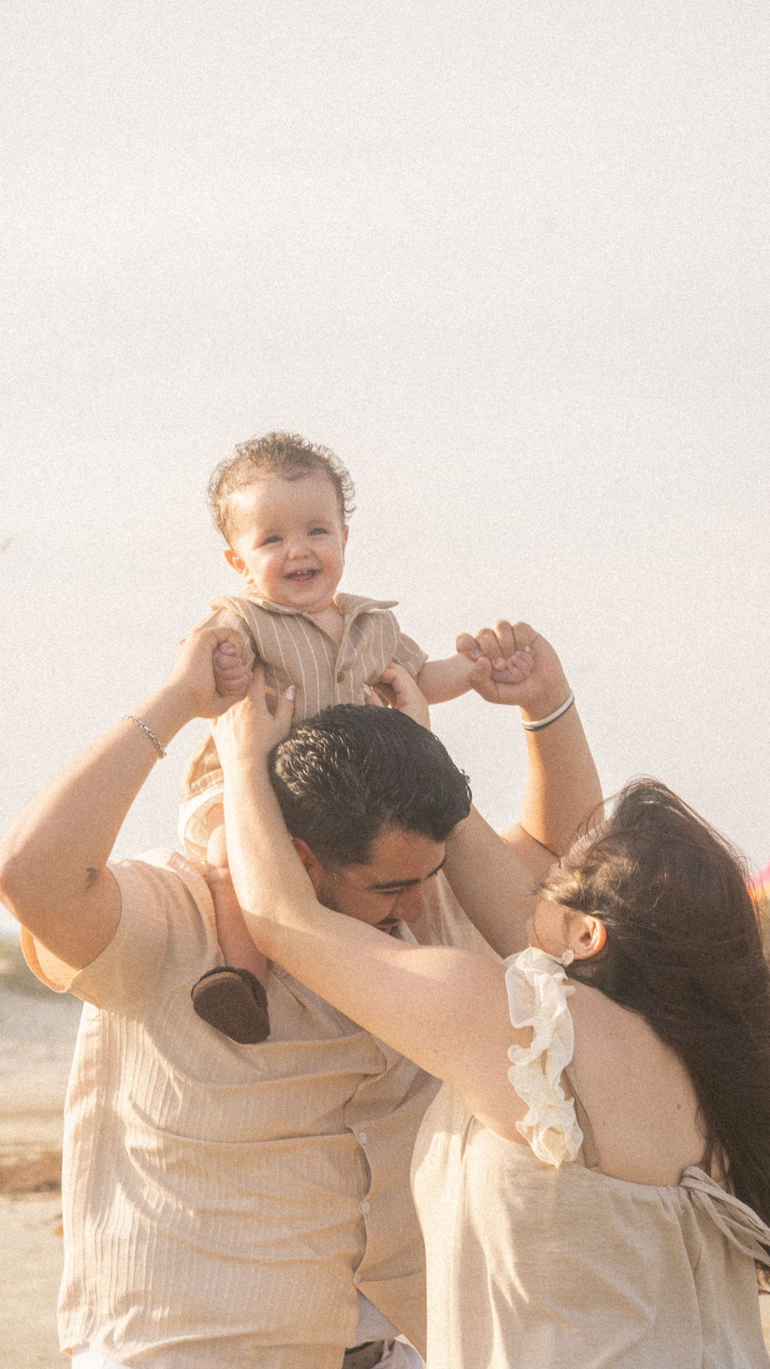 Happy baby lifted by parents on a sunny beach, all dressed in neutral tones, sharing a joyful family moment.