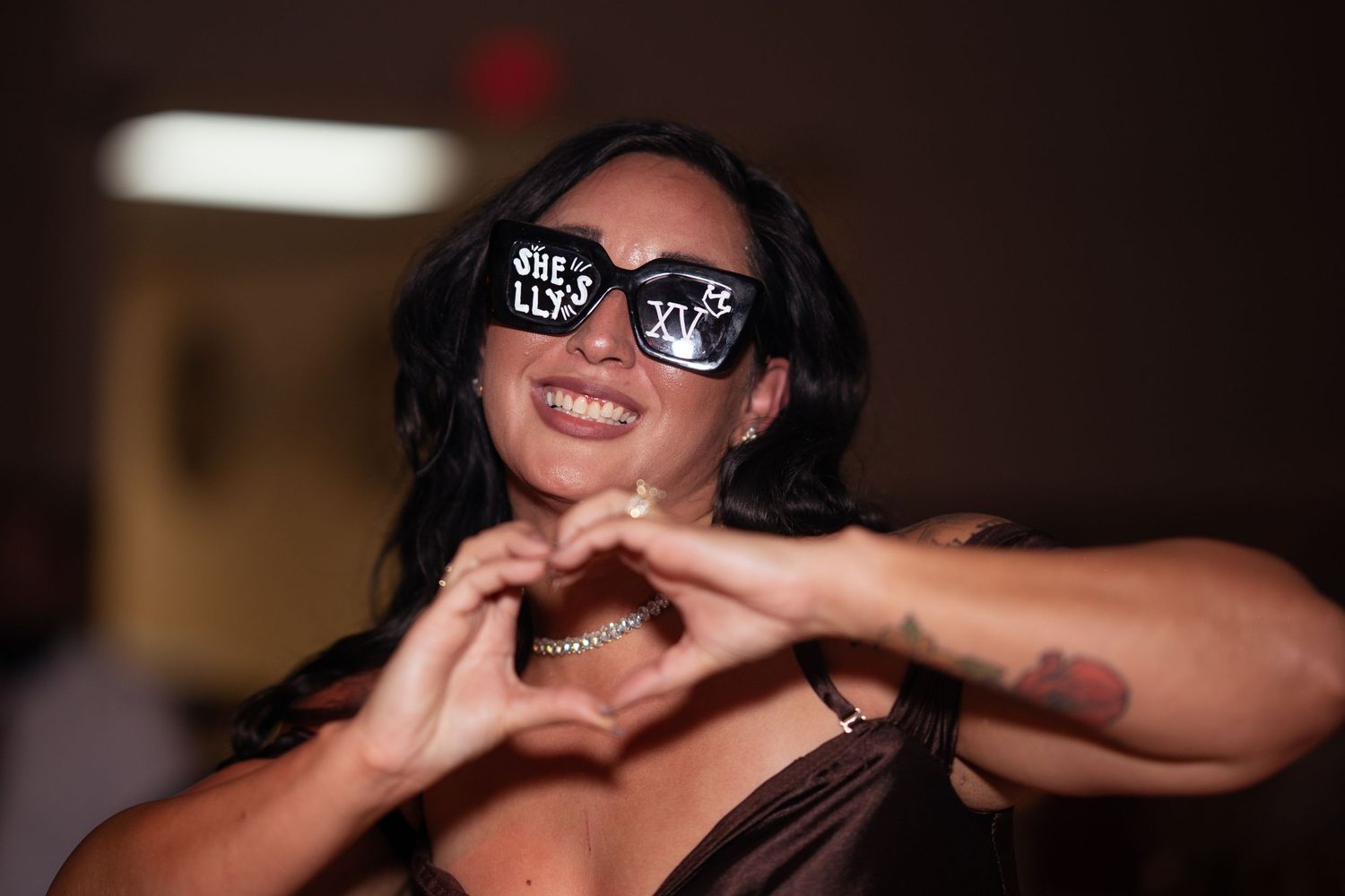 Woman smiling and making a heart shape with her hands wearing decorative quinceañera sunglasses.
