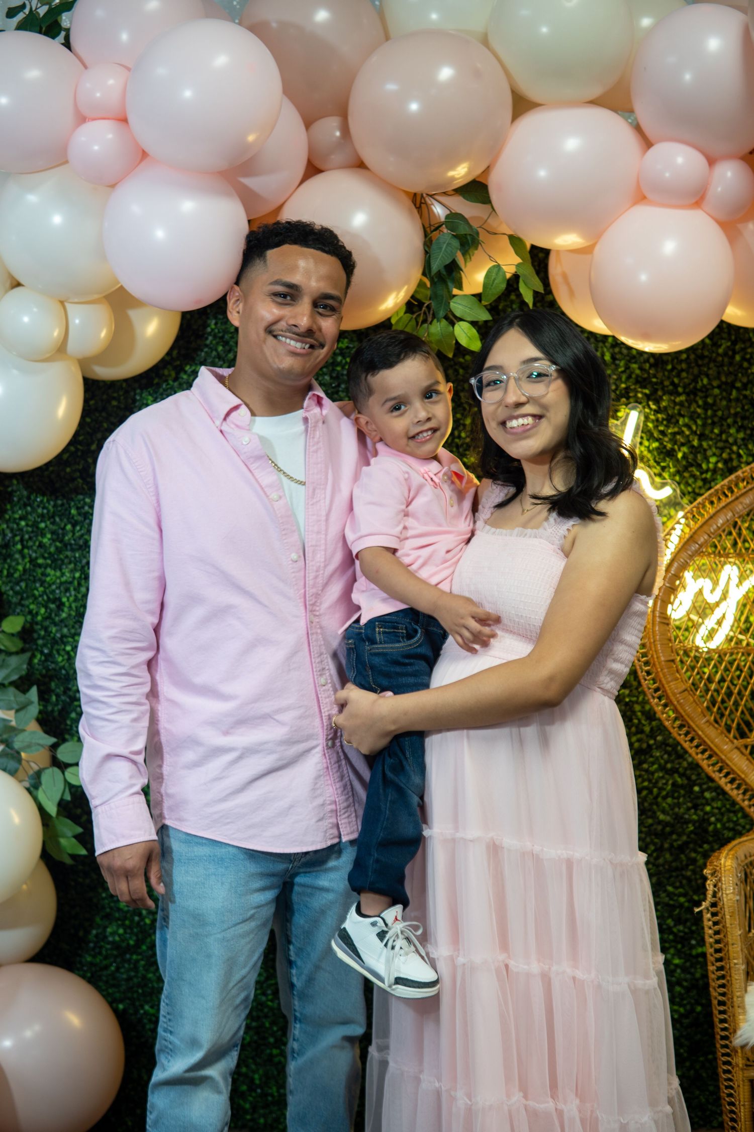 Family poses together in front of pink balloon arch and greenery backdrop at celebration party.