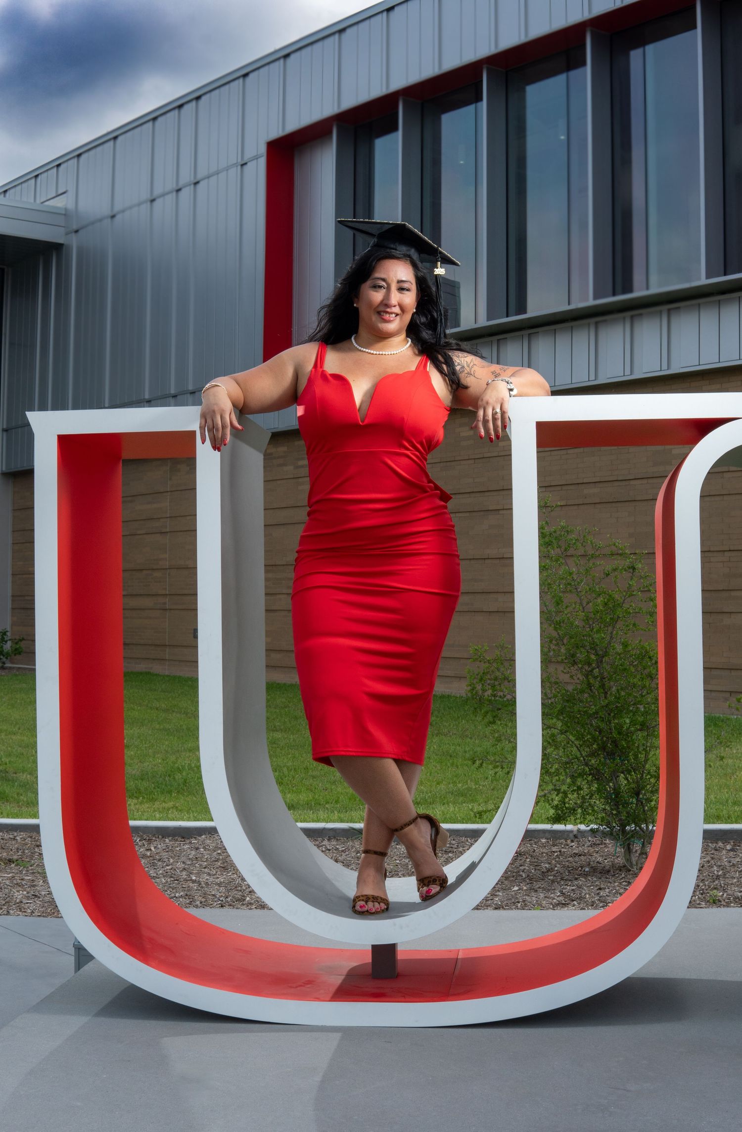 Graduate in red dress poses with large decorative U-shaped sculpture outside modern building during commencement celebration.