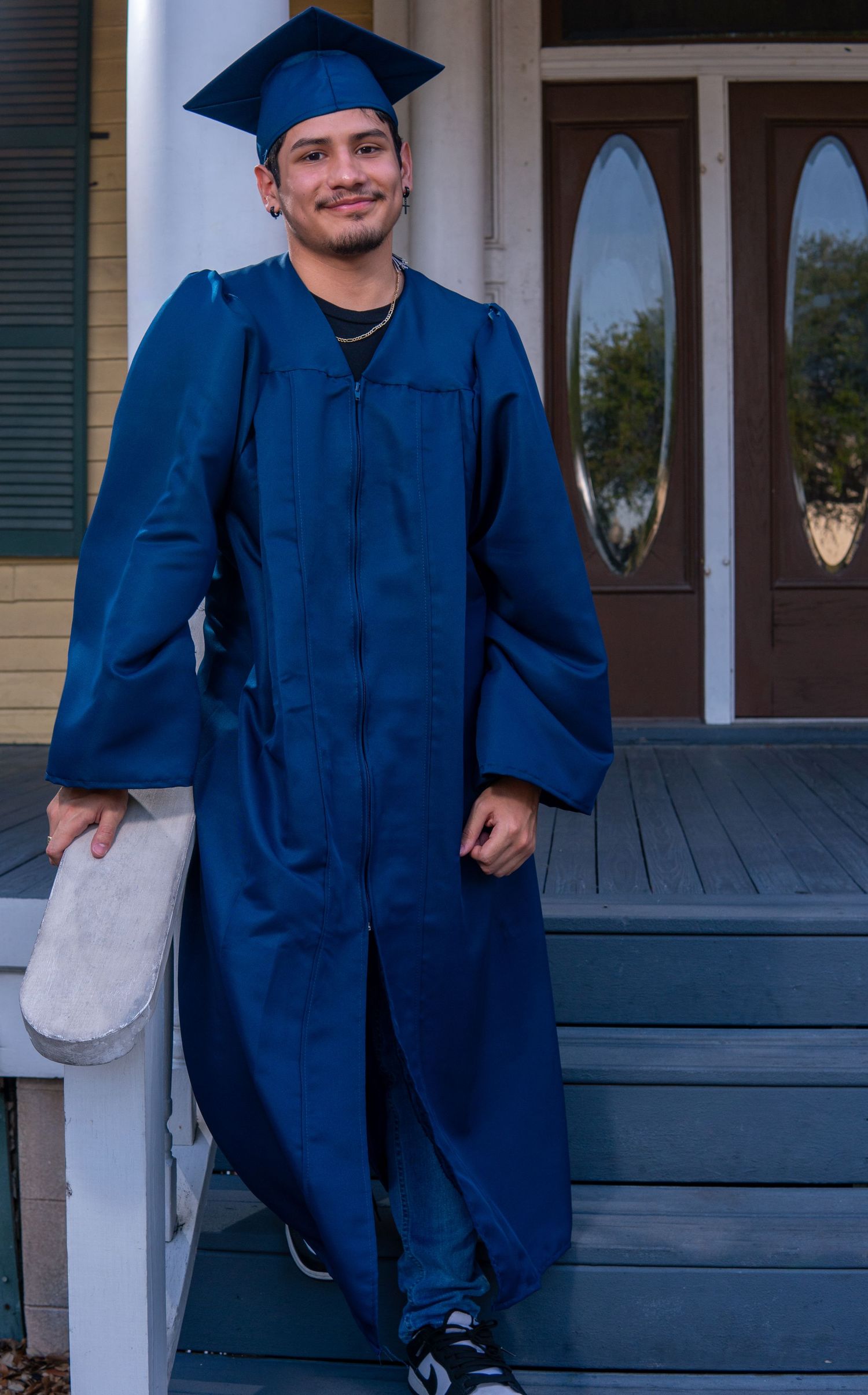 A graduate in a royal blue cap and gown stands on outdoor porch steps near a door with decorative oval windows.