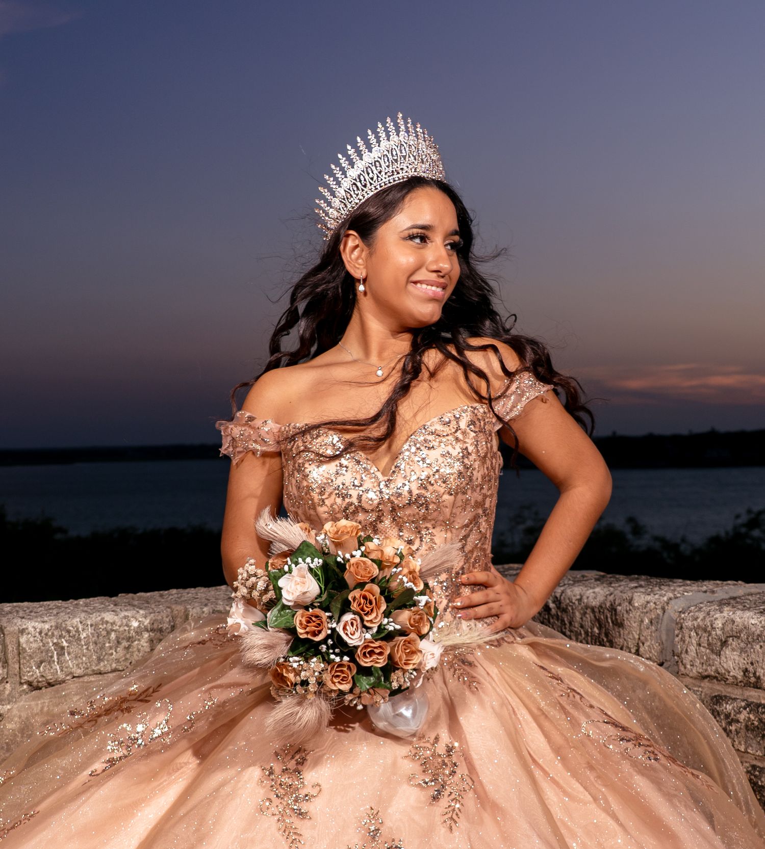 A quinceañera poses in a glittering rose gold ball gown with a tiara and bouquet during sunset by the water.