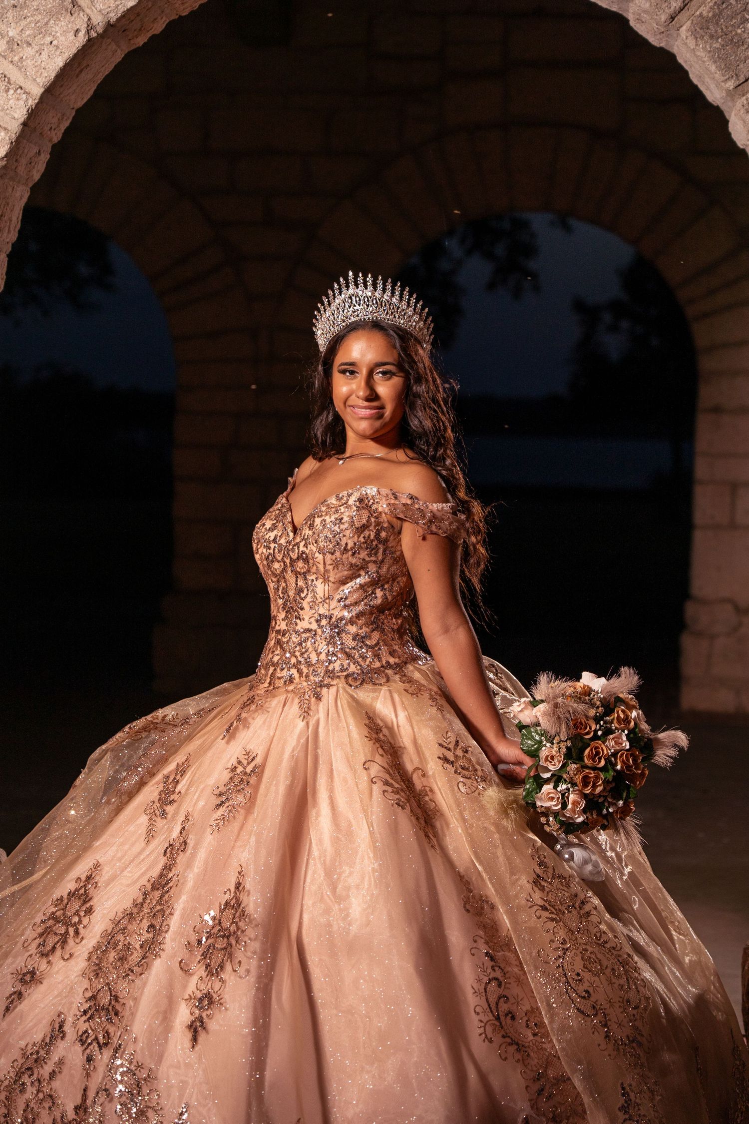 Luxurious quinceanera dress with off-shoulder design and ornate beading captured under stone archway.