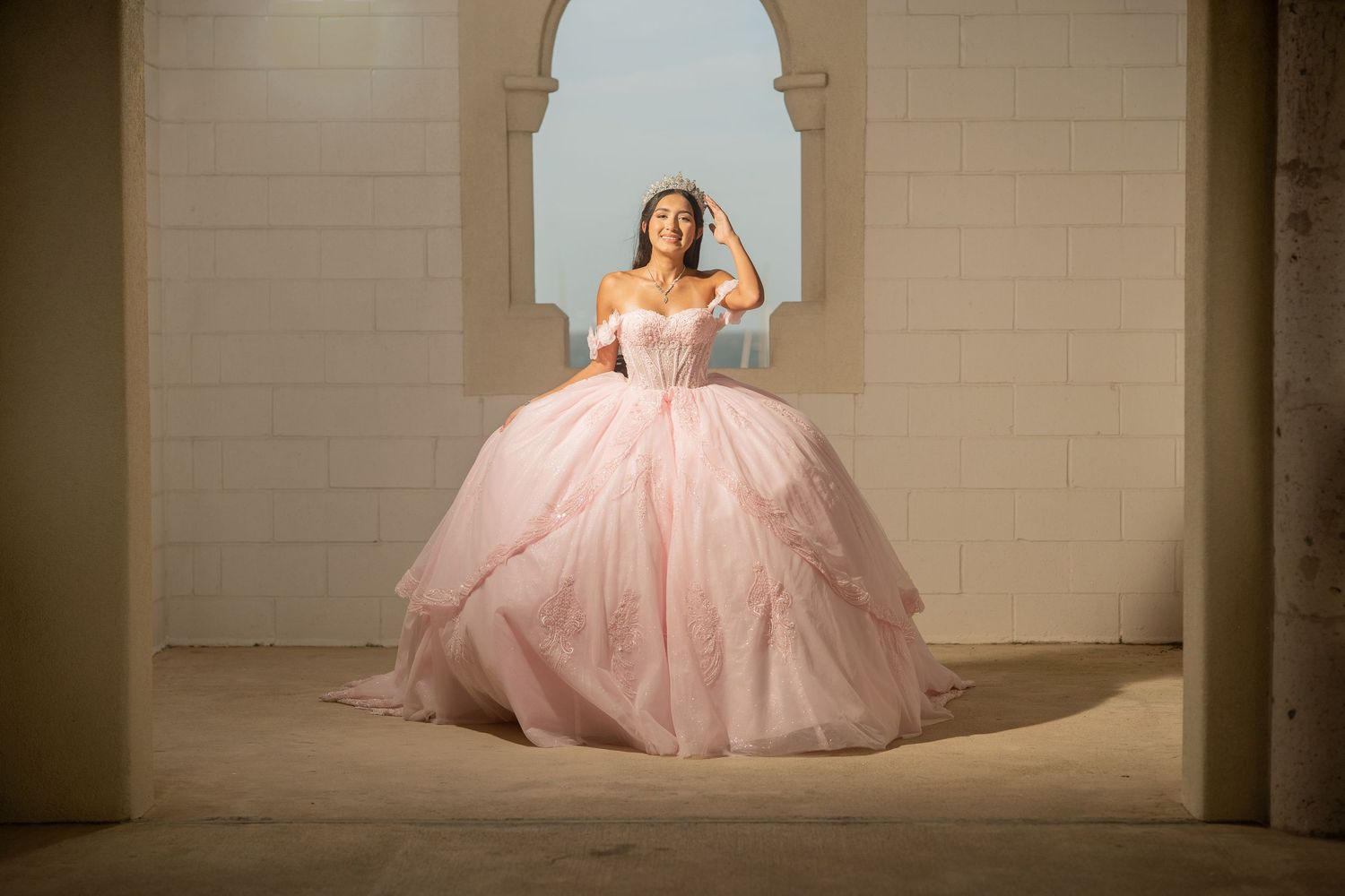 Young woman in a stunning pink ball gown quinceañera dress poses elegantly in an archway.