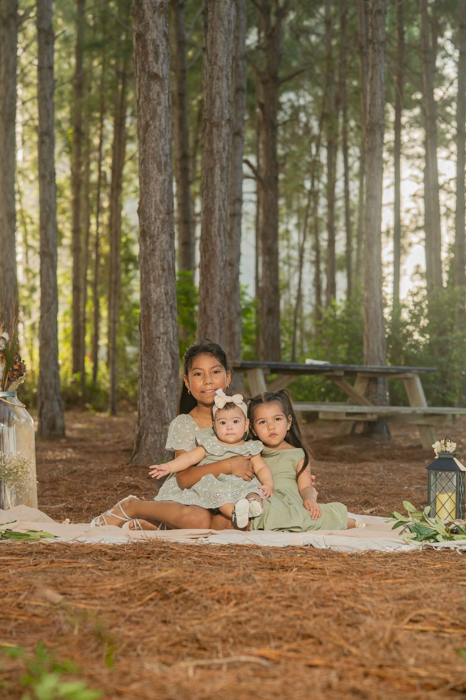 Three sisters in green dresses pose together on a blanket among tall pine trees in a serene forest setting.