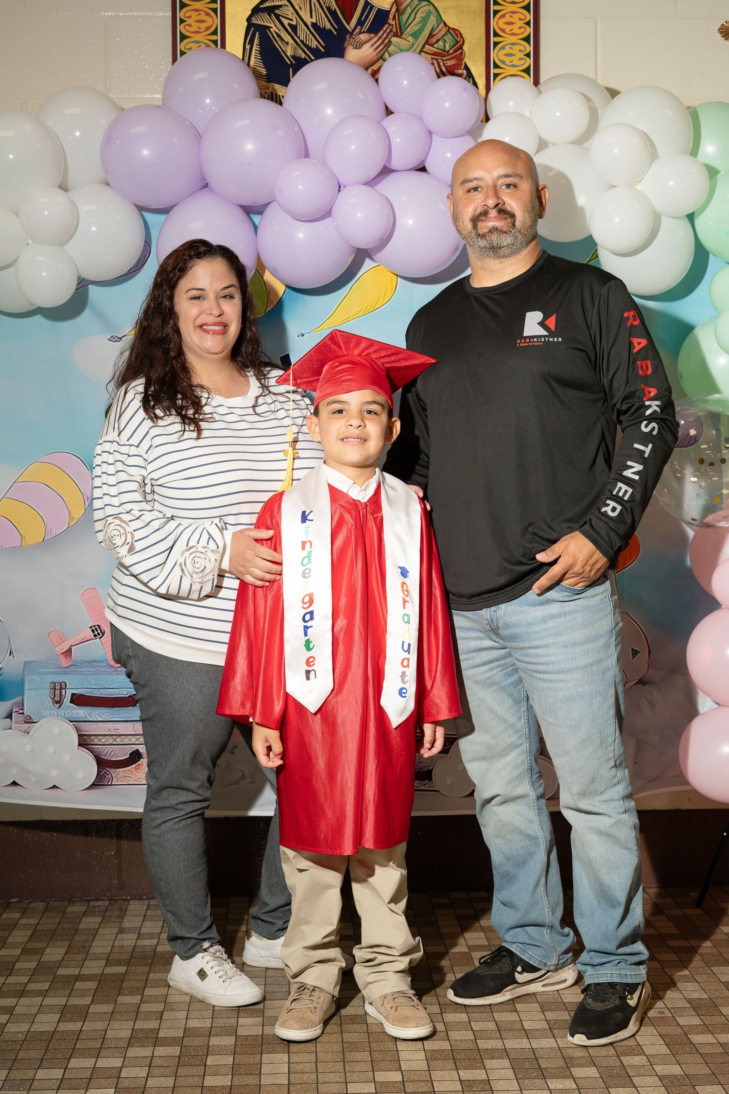Young boy in red graduation cap and gown poses with proud parents in front of balloon backdrop.