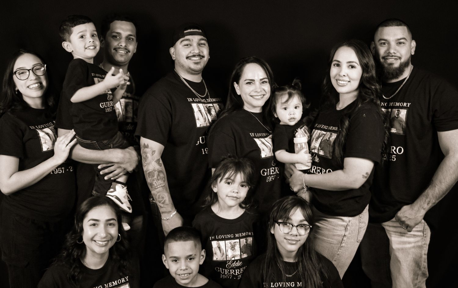 Black and white studio portrait of a large family group wearing matching t-shirts and posing together.