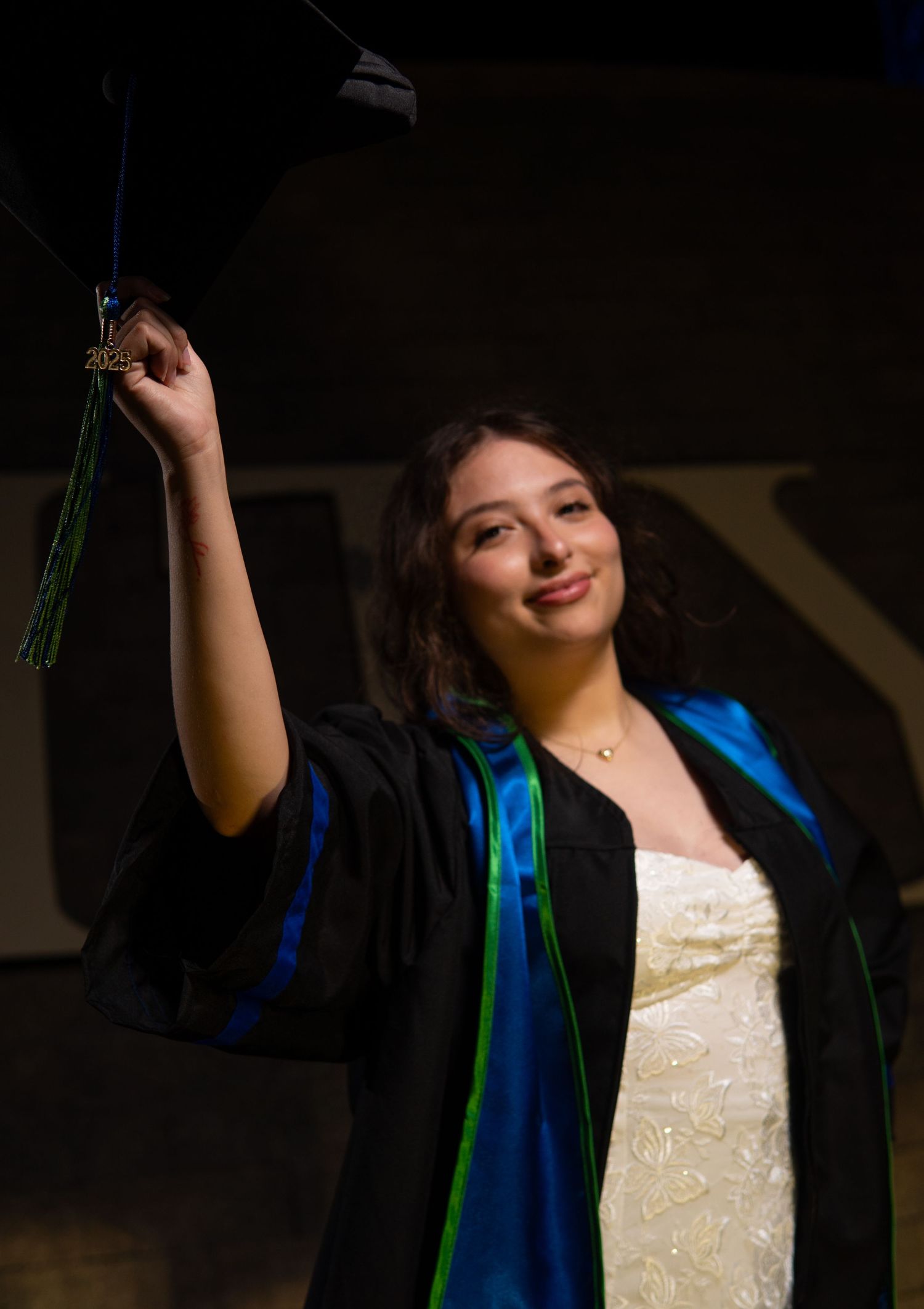 Graduate in dark academic regalia with blue sash holds up tassel against dark background in celebration.