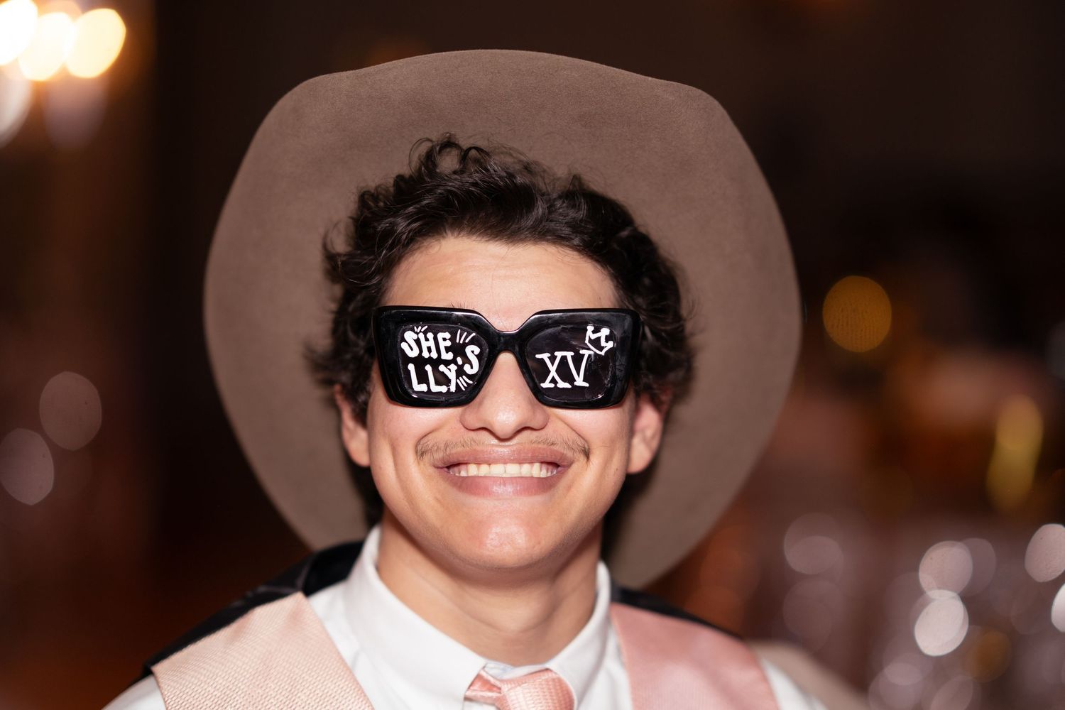 Smiling young man wearing a cowboy hat and novelty sunglasses reading XV at a quinceañera celebration.