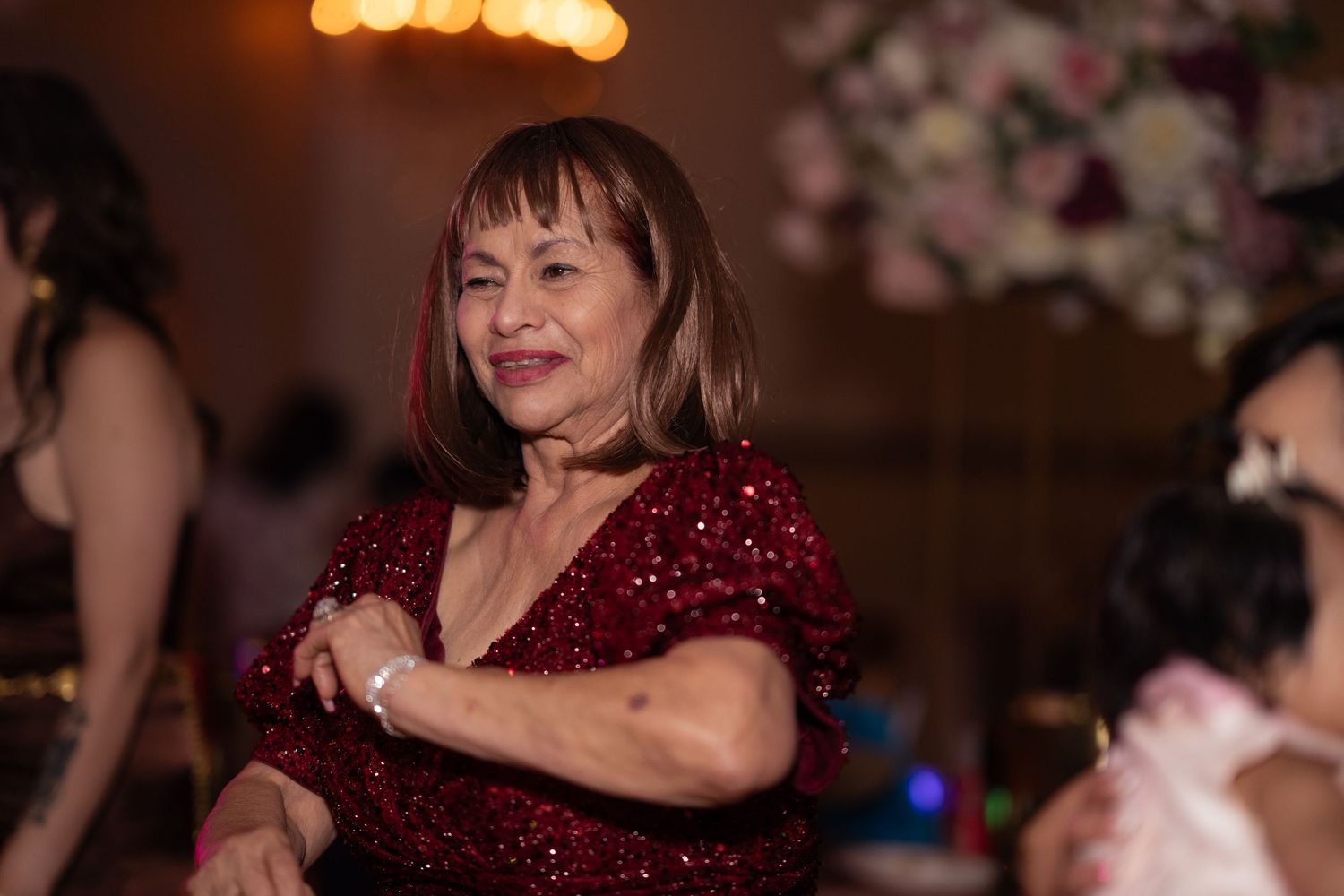 Elegant older woman in red sequin dress dancing at a formal event with floral decorations in background.