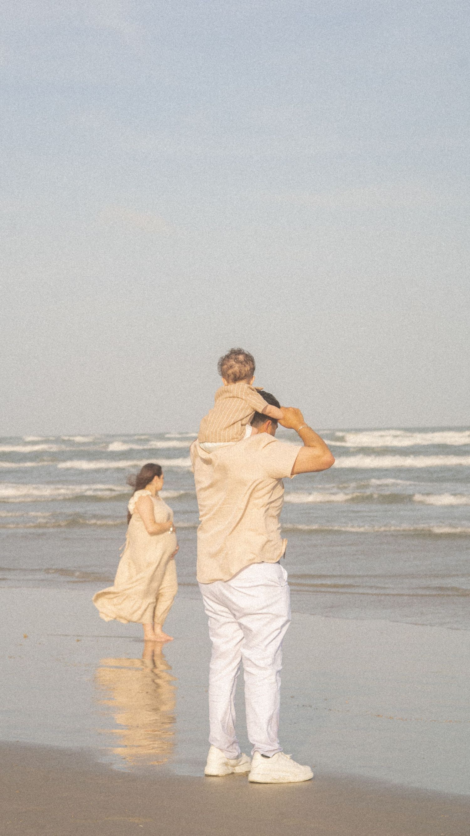 Family of three enjoying a beach day, father carrying child on shoulders while pregnant mother walks along the shoreline.