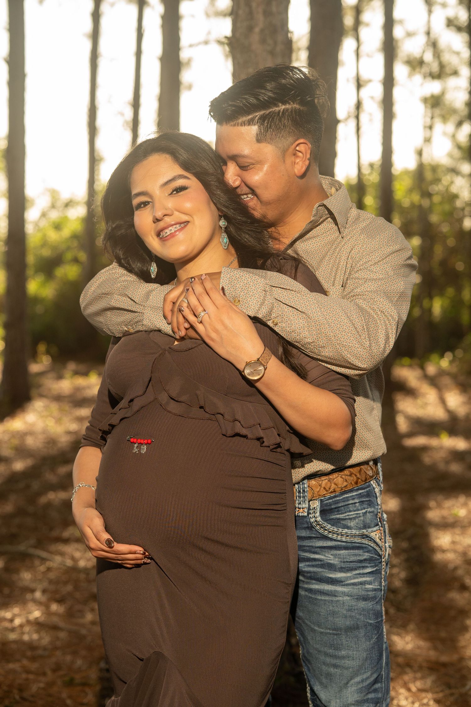 A pregnant woman in a brown dress smiles as her partner embraces her from behind in a sunlit forest.
