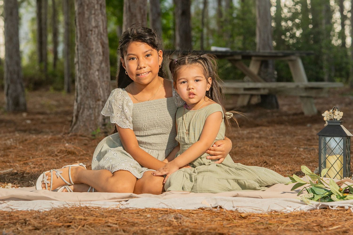 Two young girls in matching green dresses sit together on a blanket in a pine forest for a portrait photo.