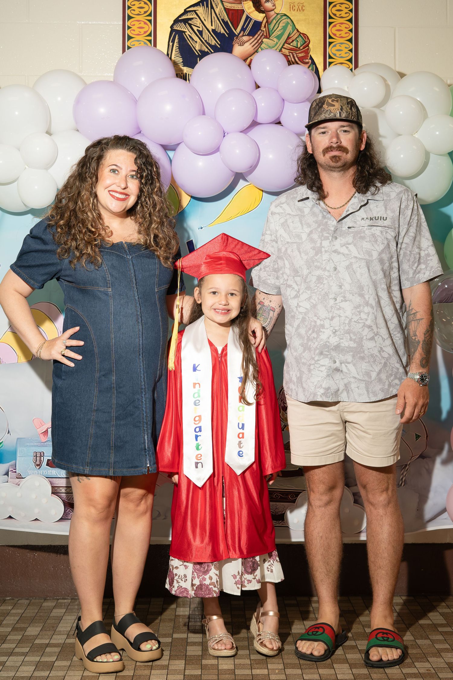 A young girl in red kindergarten graduation cap and gown poses with her proud parents in front of balloon decorations.