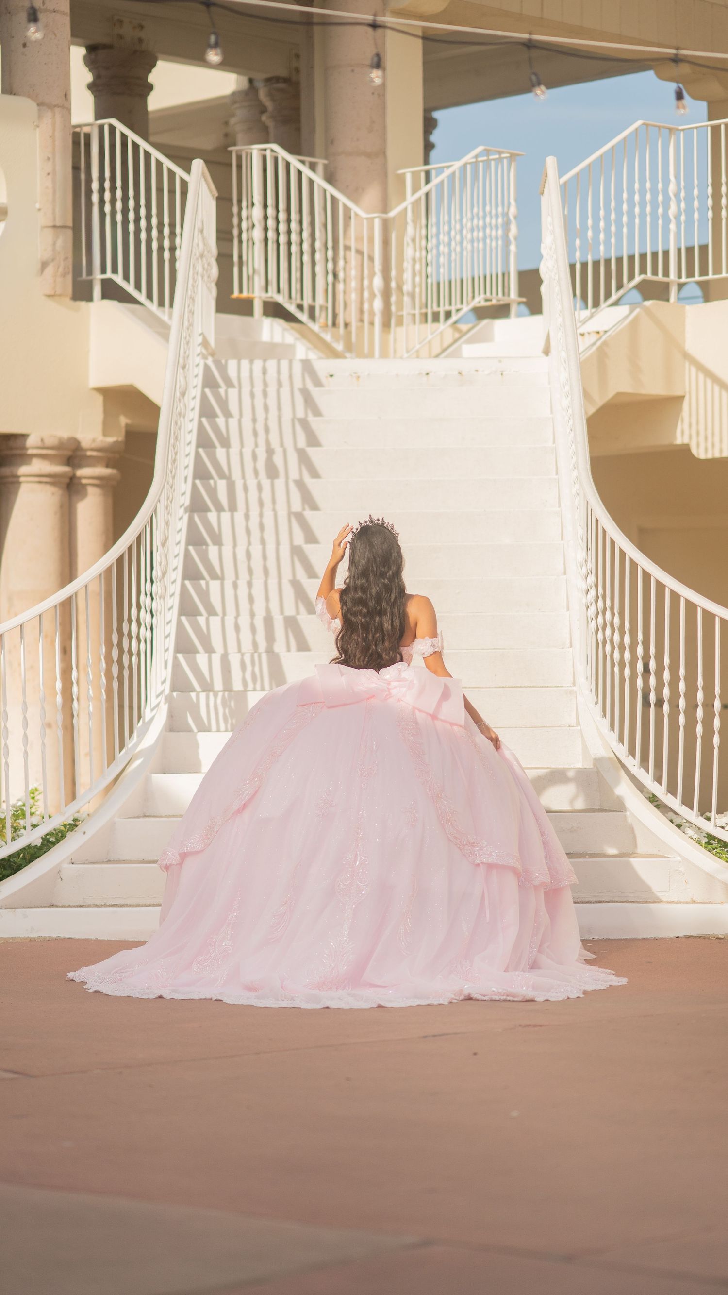 Young woman in pink ball gown and tiara poses on white staircase for quinceañera celebration.