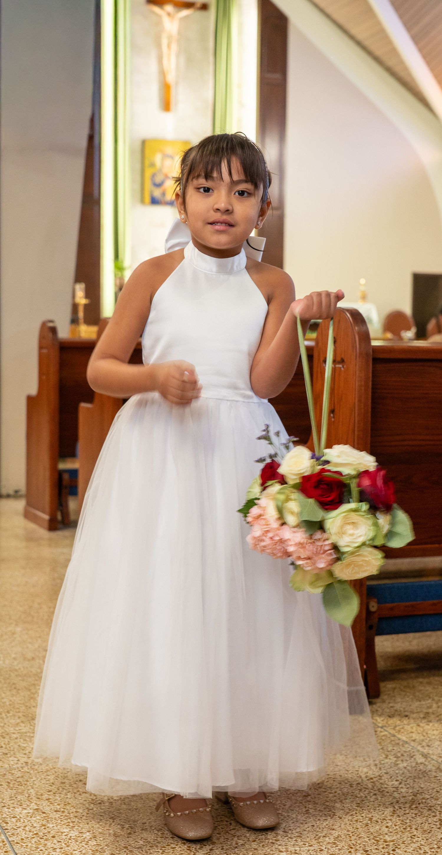 Young girl in white dress holds flower bouquet in church during First Communion ceremony.