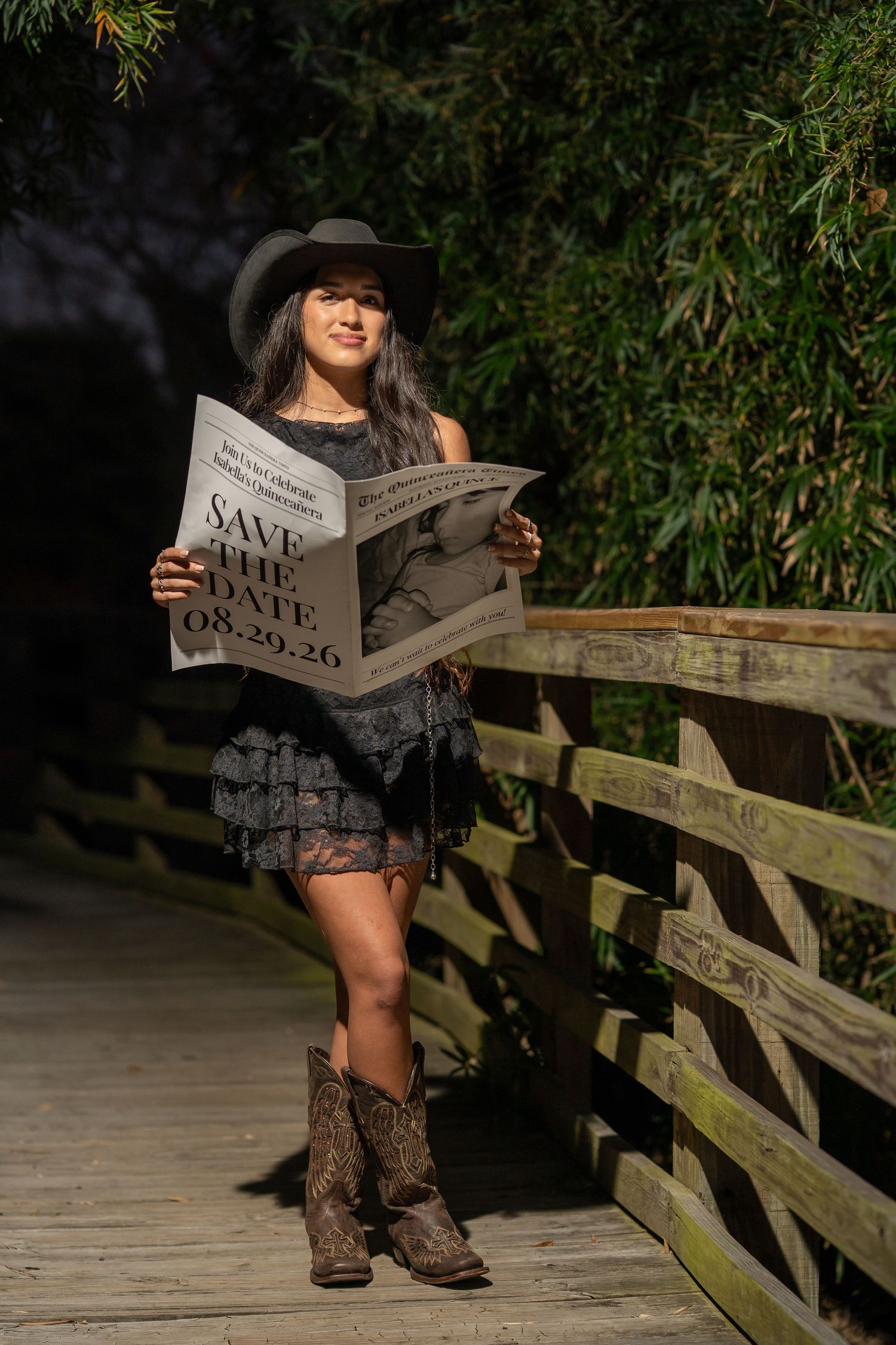Woman in black cowboy hat and boots holds Save the Date newspaper on wooden bridge surrounded by lush greenery.